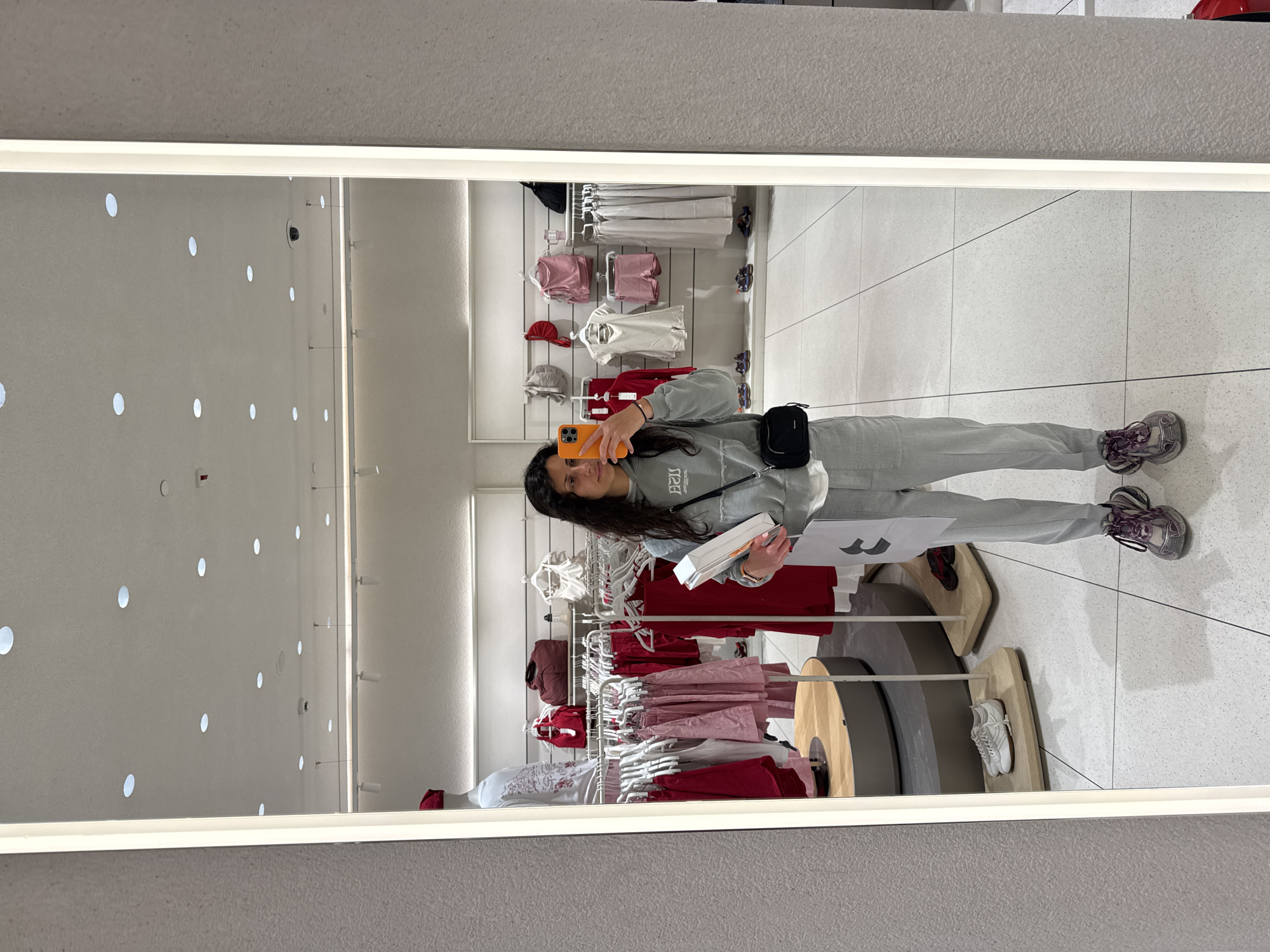 An indoor mirror selfie captures a young woman standing in what appears to be a clothing retail store. She has long, dark, wavy hair and wears a grey tracksuit, consisting of a hoodie with "1920" printed on it (reversed in the reflection) and matching sweatpants. She also wears grey and purple sneakers and a black fanny pack or crossbody bag. In her right hand, she holds an orange smartphone, presumably taking the picture, while her left hand holds a white box or package and some papers, one of which prominently displays a large black outline of a "W" or "M" logo. She has a neutral expression.
The background, visible through the mirror's reflection, features multiple clothing racks displaying various garments in white, light pink, and red/maroon hues, including t-shirts, shorts, and skirts. Accessories like hats and small bags are also seen. The floor is a light-colored tiled surface with dark grout lines. The ceiling is white with numerous round, recessed lights, and a small black security camera is visible on the upper left wall in the reflection. Several circular display stands, made of wood and grey material, are on the floor, some showcasing items like white sneakers. The overall lighting is bright and artificial, characteristic of an indoor shopping environment.