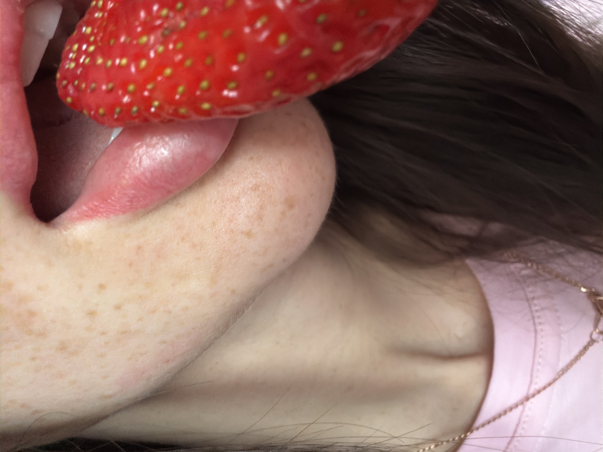 A close-up shot depicts a person with freckled skin eating a large strawberry. The strawberry is held up to the person's mouth, and their tongue is touching the fruit. The person has dark brown hair and is wearing a pink shirt. A gold necklace is visible. The location cannot be determined from the image.
