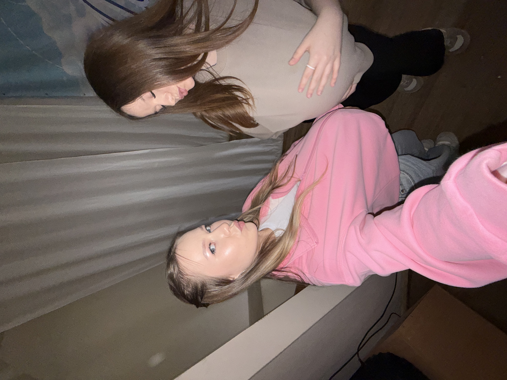 In a dimly lit indoor environment, possibly a room in Chaikovsky, Russia, two young women are captured in a selfie. The woman on the left, with blonde hair pulled back and a pink jacket over a white shirt and grey jeans, is looking at the camera with her lips pursed. The woman on the right, with long brown hair, is wearing a beige t-shirt and black leggings. She is turned away from the camera, looking down and placing her hand on her stomach. The background shows white curtains and a hint of a blue mural. The lighting suggests it might be evening or indoors with artificial light.