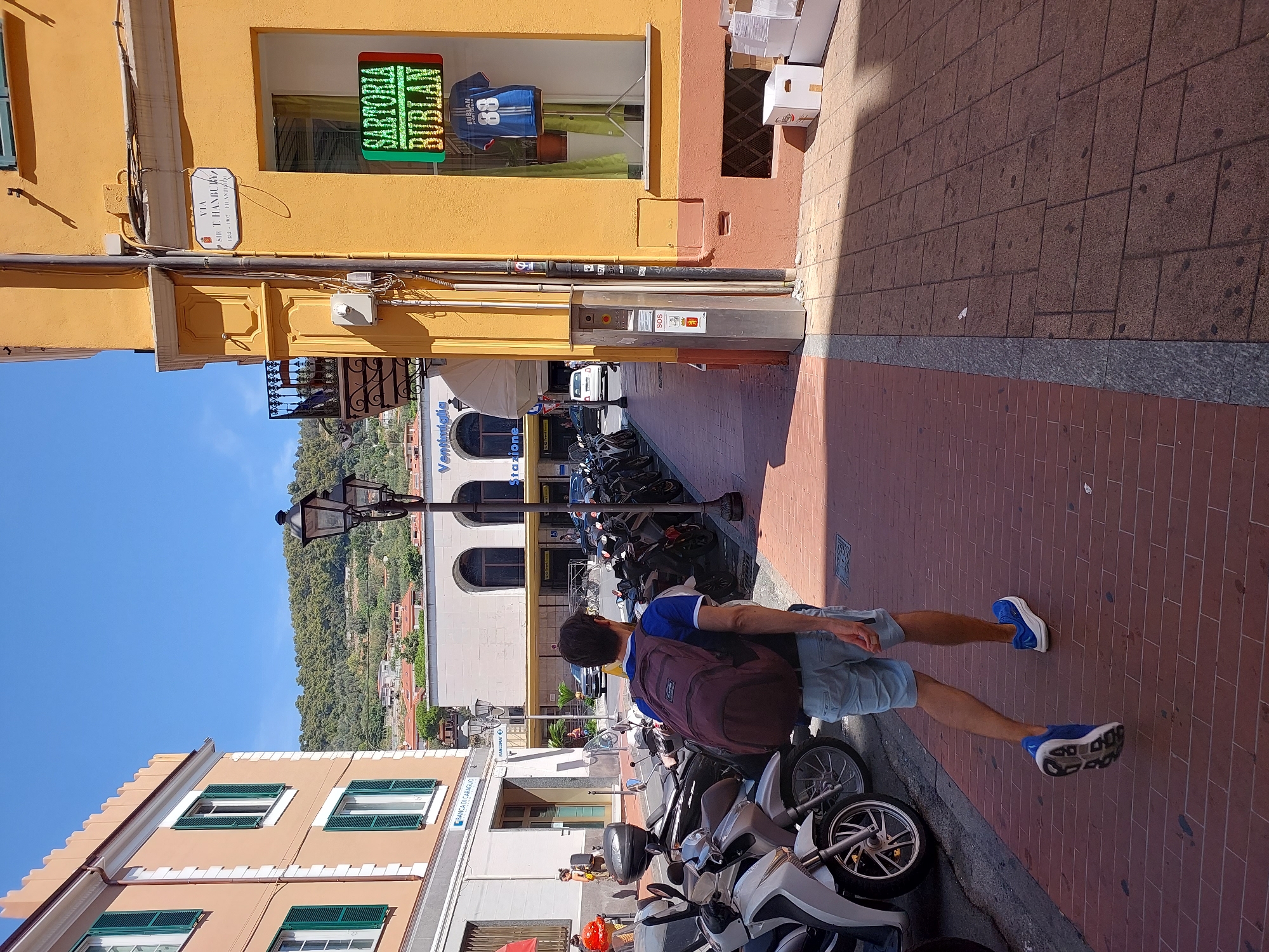 A man with a maroon backpack is walking away from the camera down a brick-paved sidewalk lined with parked scooters and motorcycles. He is wearing light blue shorts and blue athletic shoes. He is walking past a yellow building with a storefront window displaying a blue jersey with the number 88. Above the window is a street sign that reads "VIA SIR T. HANBURY 1832-1907". To the left of the yellow building is a white building with an arched entrance labeled "Stazione". The scene appears to be in Ventimiglia, Italy. The sky is clear and blue, and the sun is casting shadows. There are other people and vehicles visible in the background.