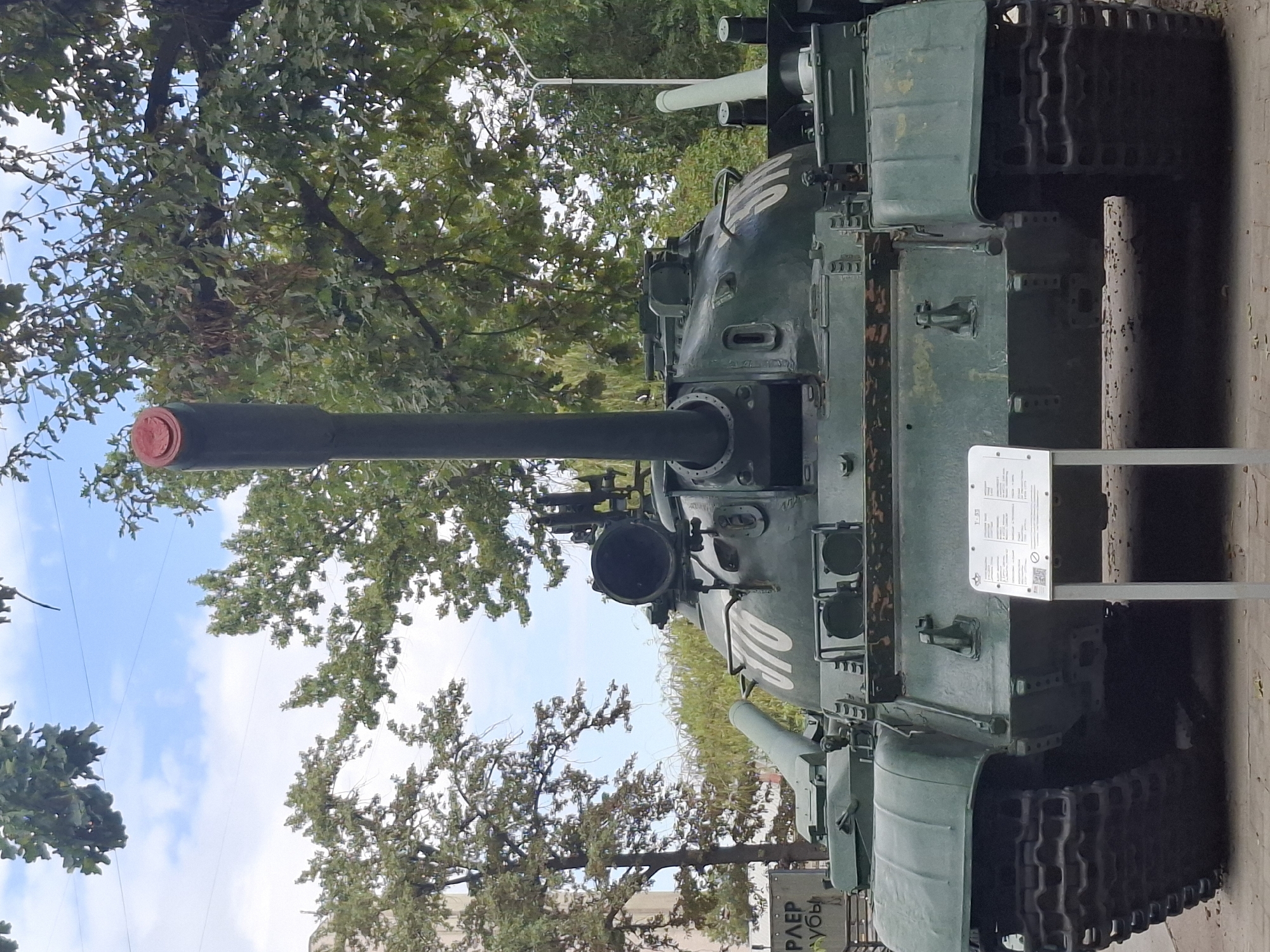 This is a medium shot, eye-level photograph of a green military tank displayed outdoors. The tank is the primary subject, positioned in the center of the frame and facing forward. Its large cannon is prominent, topped with a red cap. The tank has markings, including white numbers "070" on its turret. There is a white informational plaque in front of the tank.
The tank is situated on a paved surface. Behind the tank, a backdrop of lush green trees with leafy branches creates a natural setting. The sky above is visible, featuring scattered white clouds against a blue background. A pole with a light fixture is partially visible to the right of the tank. To the left, a portion of a building and a sign with Cyrillic text are visible.
The photograph appears to have been taken in Almaty, Kazakhstan. The presence of the tank on display suggests it is likely a monument or part of a museum. No people are visible in the image.