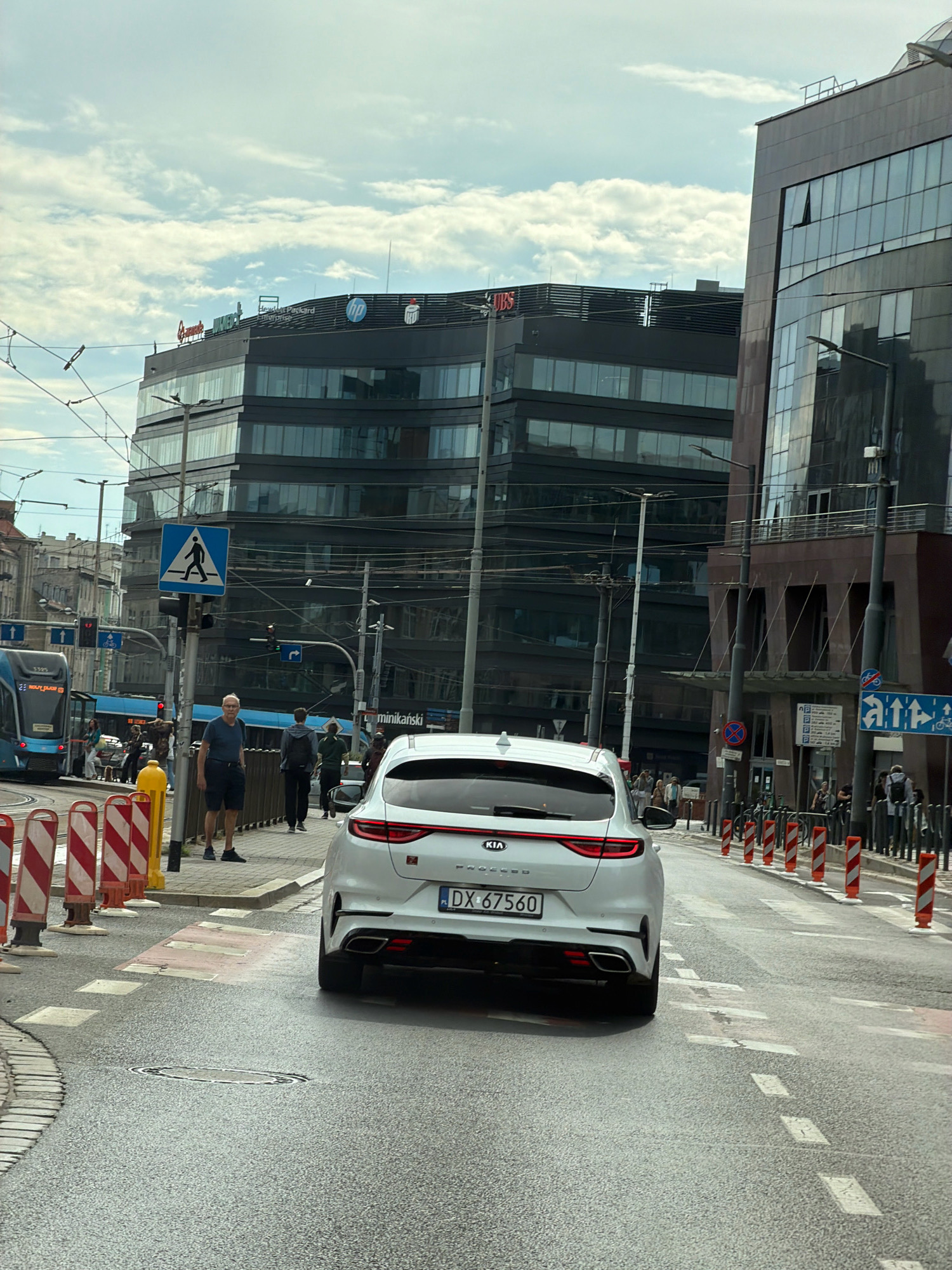 The image captures a view from inside a vehicle looking forward, at a city street in Wrocław, Poland. A white Kia hatchback with the license plate DX 67560 is in the foreground, partially obscuring a pedestrian crossing. The street is wet, suggesting recent rain. In the background, a modern dark glass building with a curved facade is visible, with another building to its right which is brown and has a similar architectural style. Several streetlights and overhead power lines crisscross the sky. A blue tram is stopped on the left side of the road, with passengers visible. A pedestrian crossing sign showing a person walking is mounted on a traffic light pole. Several people are walking on the sidewalk. Red and white striped bollards line the right side of the road. The sky is overcast with some white clouds.