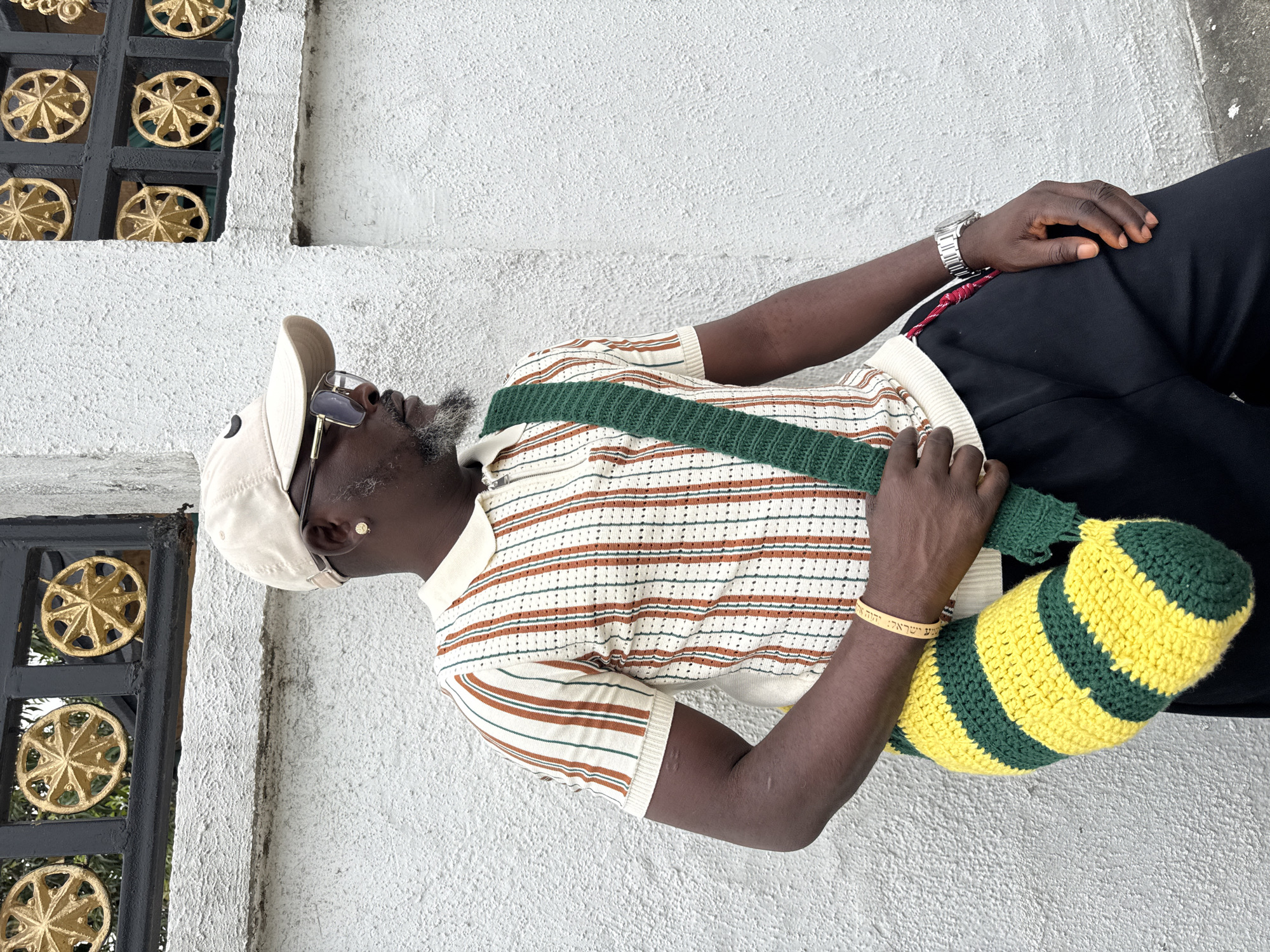 A full-body shot of a middle-aged Black man standing in Ado, Nigeria. He faces left, wearing a light-colored baseball cap, sunglasses, a short-sleeved striped polo shirt with brown and white stripes, black pants, and a silver wristwatch on his left wrist. He has a green knitted strap across his chest, attached to a yellow and green striped knitted bag. On his right wrist, he has a gold bracelet with Hebrew lettering. He stands against a textured white wall with black metal latticework featuring gold rosette designs.