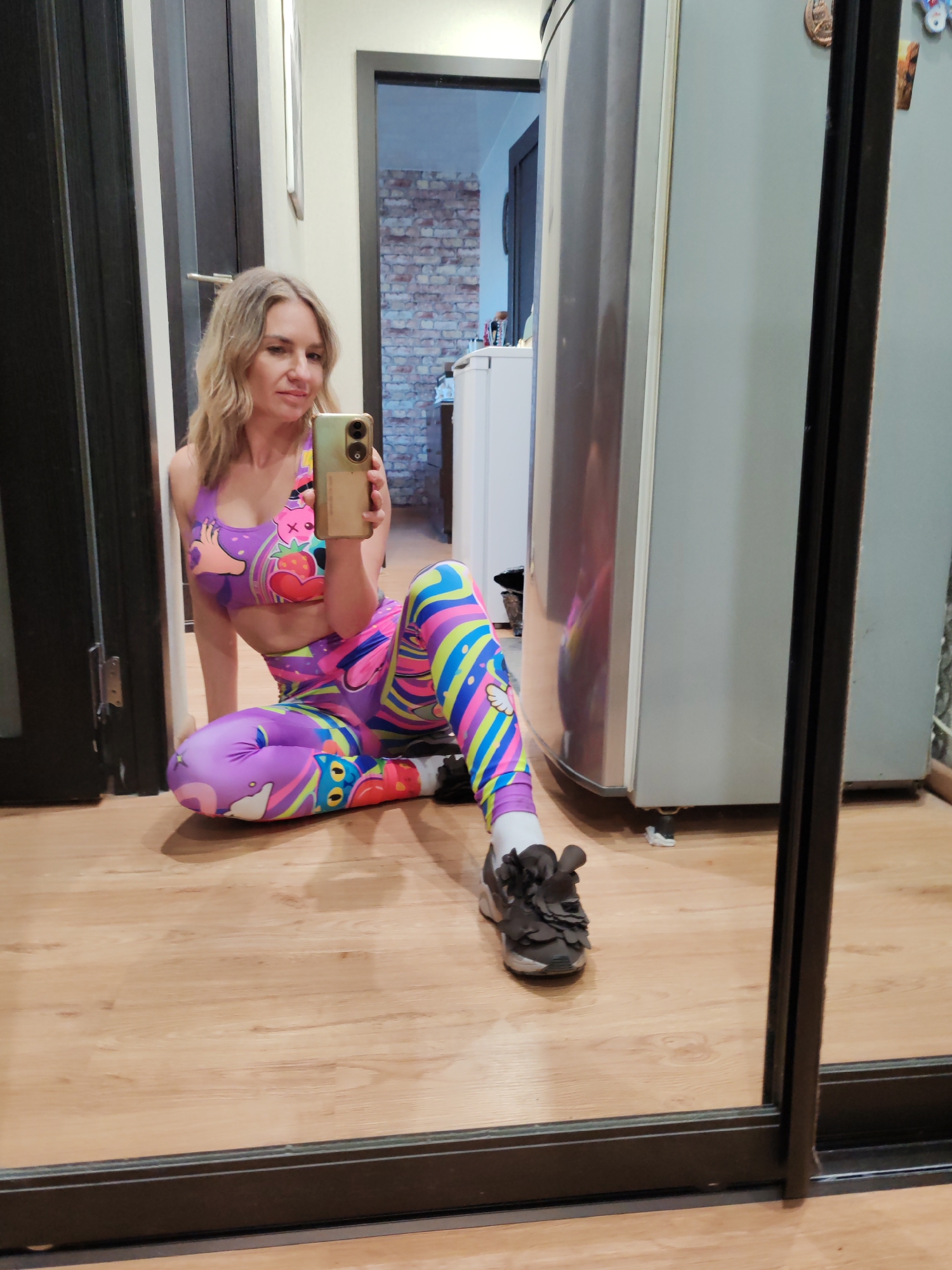 A woman is sitting on a wooden floor in an indoor setting, taking a selfie with her smartphone. She is wearing a colorful patterned sports bra and leggings. One shoe is visible on her foot, a dark-colored sneaker with floral embellishments. A refrigerator is to her right, partially obscured by a sliding mirror door. Behind her, a doorway leads into another room with a brick-patterned wall and a white cabinet. The location is likely a residence, and there is no evidence indicating the city of Petropavlovsk-Kamchatsky, Russia.