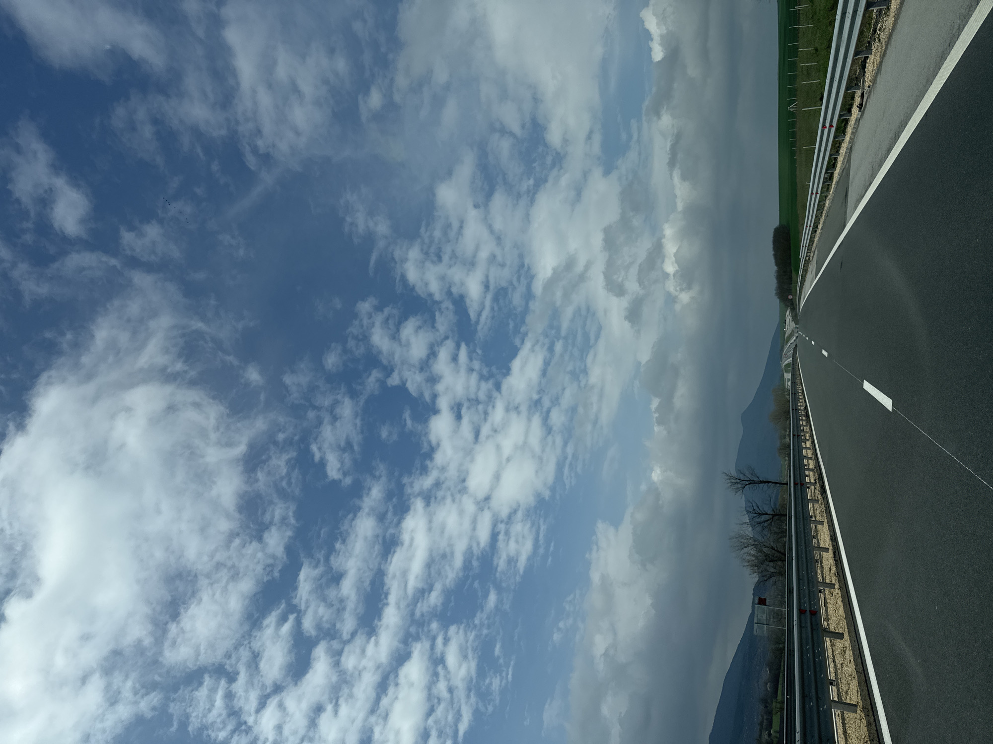 The image depicts a highway in Gradishte, Bulgaria, under a partly cloudy sky. The road is asphalt, divided by white lines. To the right of the highway, there's a metal guardrail and a grassy field. In the distance, mountains are visible through the clouds.