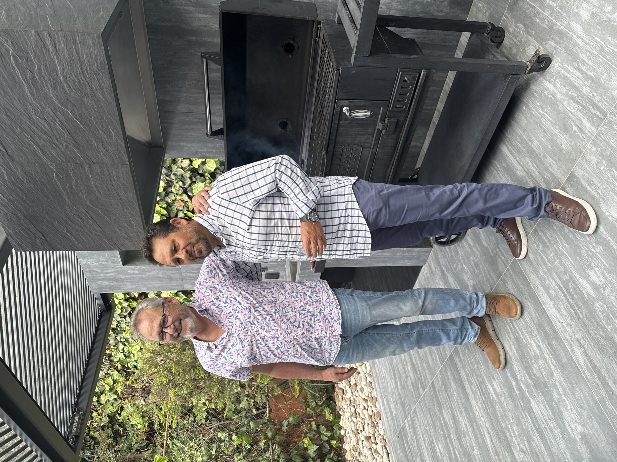 Two men are captured in an outdoor setting, standing next to a large black barbecue grill or smoker. The image shows a relaxed, daytime gathering.
The man on the left, appearing older, wears glasses and has a grey beard. He is dressed in a short-sleeved shirt with a pink and blue abstract pattern and blue jeans, complemented by brown shoes. He stands with a faint smile, looking towards the camera.
The man on the right, younger and also bearded, has his arm around the older man's shoulder. He is wearing a white and blue checkered long-sleeved shirt with rolled-up sleeves, dark blue pants, and brown shoes. He also looks towards the camera with a subtle smile, indicating a friendly interaction.
Behind the men, a substantial black barbecue grill/smoker is visible, from which a wisp of smoke rises, suggesting it is in use. The brand name "SMOKE HOLLOW" is legible on its side. The grill is equipped with wheels, making it portable.
The ground is paved with large, textured, light gray tiles, contributing to a modern aesthetic. In the background, there's dense green foliage, primarily ivy, creating a natural backdrop. Parts of the background also show light-colored pebbles or small stones. A dark, slatted architectural element is visible on the far left.
The overall scene conveys a casual and social event, likely a barbecue, taking place in an outdoor patio or terrace area. The bright lighting suggests it's during the day. The setting appears to be a well-maintained leisure space, possibly in Huixquilucan de Degollado, Mexico. Both men display relaxed and friendly expressions.