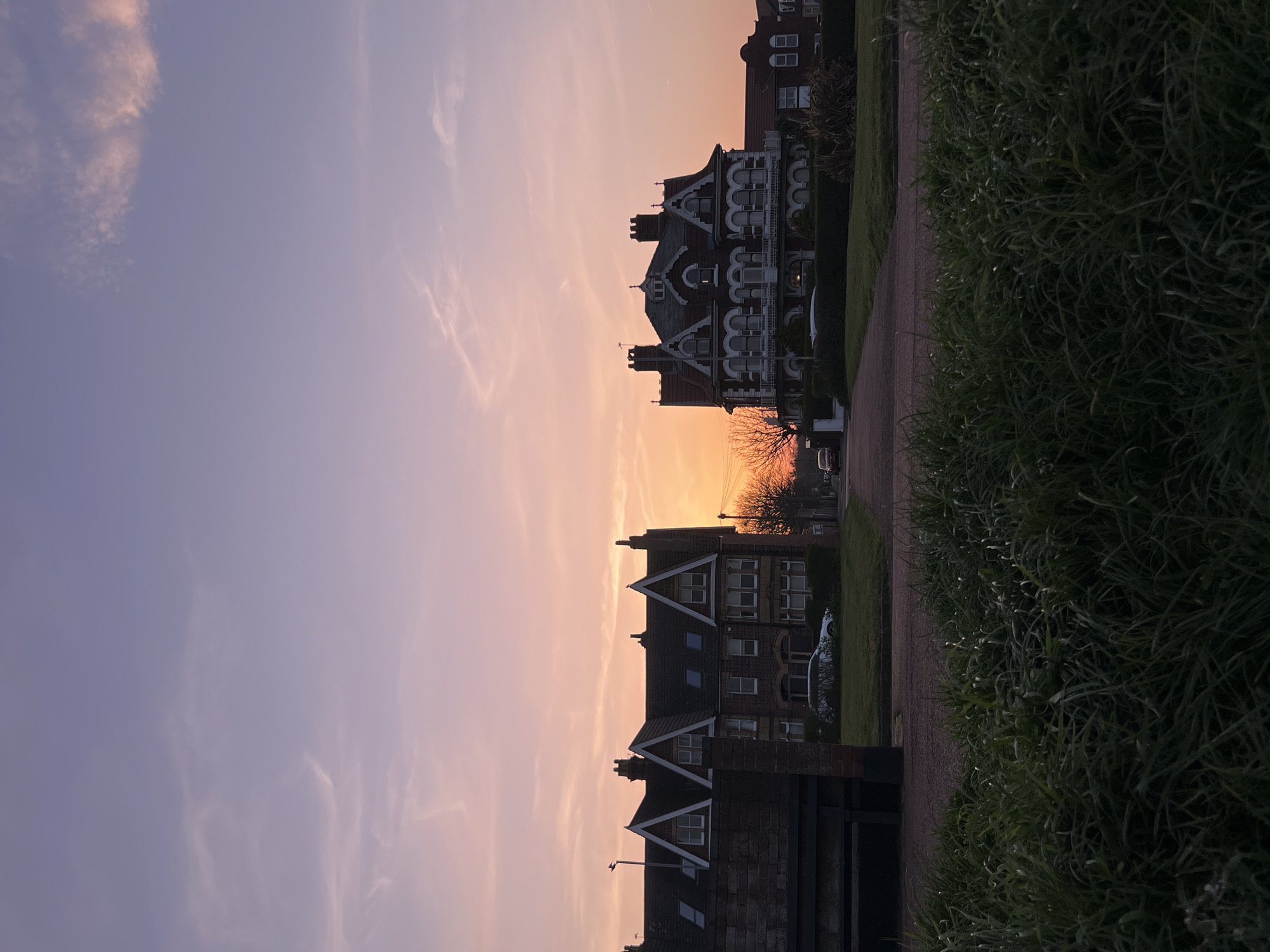 The image depicts a row of buildings in Gorleston-on-Sea, United Kingdom, taken during sunset. The sky transitions from purple to orange as the sun sets behind the buildings. The architecture is mostly brick, with some buildings having white architectural details. There's vegetation growing alongside the buildings and the road/sidewalk, suggesting a residential area. The image appears to have been taken from street level, with the viewpoint at an angle looking slightly up at the buildings.