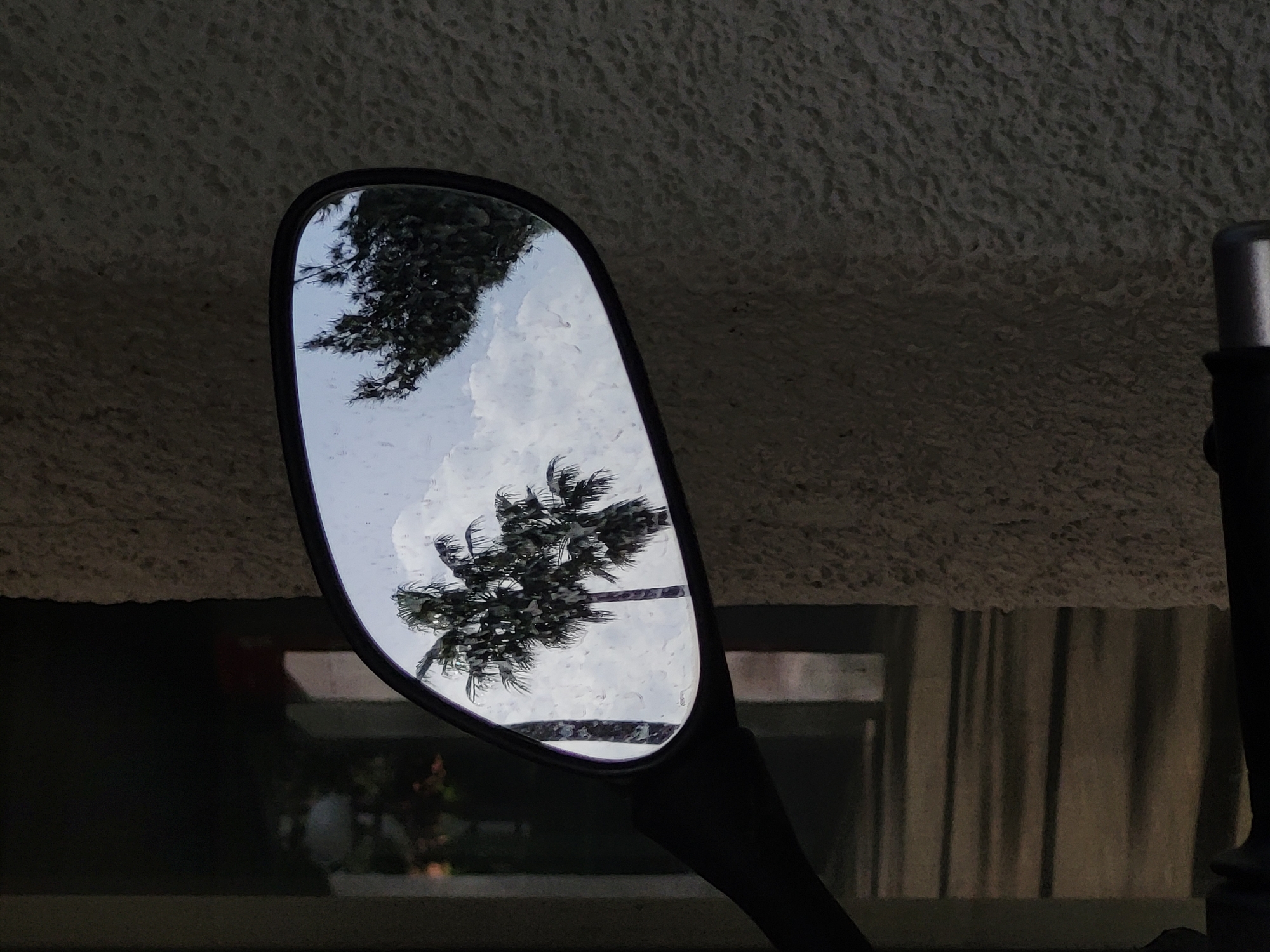 The image shows a motorcycle side mirror reflecting a cloudy sky and palm trees. Raindrops are visible on the mirror's surface. The background behind the mirror reveals part of a building with a textured wall and a window with curtains. It is not possible to confirm from the image alone that the location is in Budva, Montenegro.