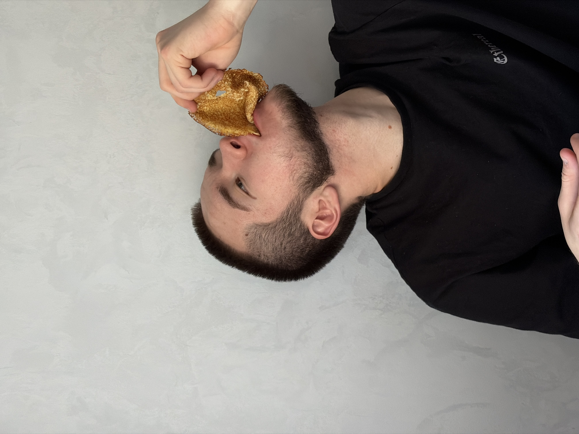 A man with a beard is eating a pancake. He is wearing a black t-shirt and is oriented vertically. The background is a textured grey wall. There is no information to determine the location as Sarapul, Russia.