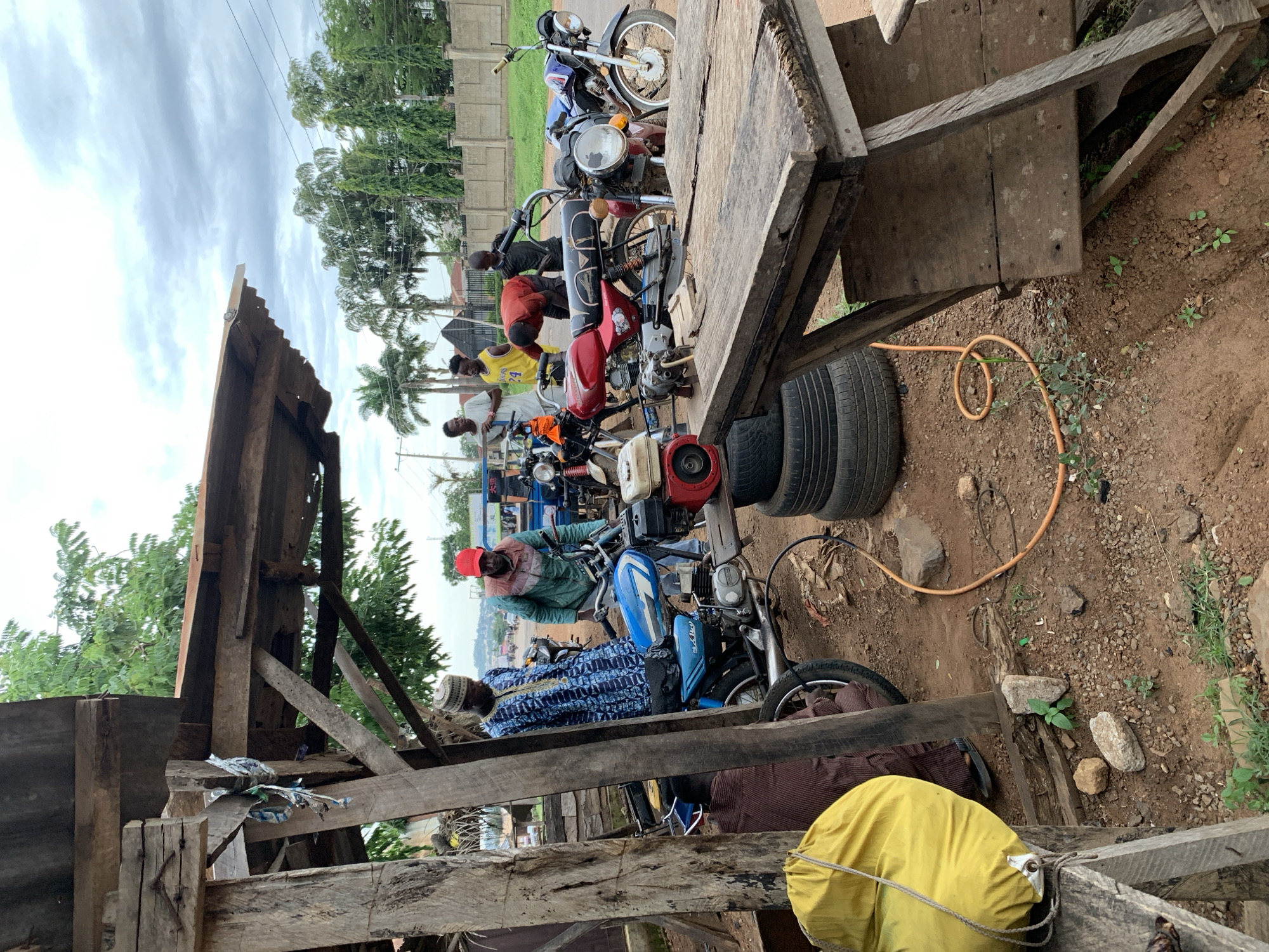 This image captures an informal outdoor motorcycle repair or service area in Gbeleko, Nigeria. The scene is set on reddish-brown dirt and gravel ground, interspersed with small rocks and sparse green vegetation, under a predominantly cloudy sky.
In the foreground on the left, a yellow cylindrical bag or sack with a rope drawstring is visible, resting on a wooden surface. Below and behind this, a person is partially obscured by vertical wooden support beams, wearing dark trousers and sandals, indicating a seated or crouching posture. On the right foreground, a rustic wooden table or workbench, constructed from rough planks, partially obstructs the view of the midground.
The midground features seven males and four motorcycles. From the left:
* A man wearing a white cap (kufi) and a blue, long-sleeved garment with white patterns stands, looking down towards a light blue motorcycle.
* Adjacent to him, a man wearing a red baseball cap, a green and brown jacket, and light trousers is positioned near the handlebars of the light blue motorcycle, which has its engine exposed.
* On the ground next to the blue motorcycle, a small engine unit with red and white components is visible. Another identical small engine unit rests on a stack of three black vehicle tires. An orange hose or cable extends from this area, coiling on the ground towards the right.
* Further right, two men stand upright, observing: one in a white shirt and the other in a yellow basketball jersey featuring the number "24."
* A man in a red t-shirt is bent over a red motorcycle, centrally positioned, appearing to work on its engine area.
* To the right of the red motorcycle, a younger male in a dark t-shirt stands near its handlebars.
* A dark-colored motorcycle (possibly purple) is parked to the far right of this group.
A simple, open-sided wooden shelter structure, comprising wooden posts, beams, and a corrugated metal roof, partially covers the left and upper parts of the scene.
In the background, a paved road runs horizontally across the image, with distant vehicles and structures visible. A low concrete perimeter wall with square pillars delineates the property line, behind which numerous green palm trees and other dense foliage grow, extending towards the horizon under the cloudy sky.