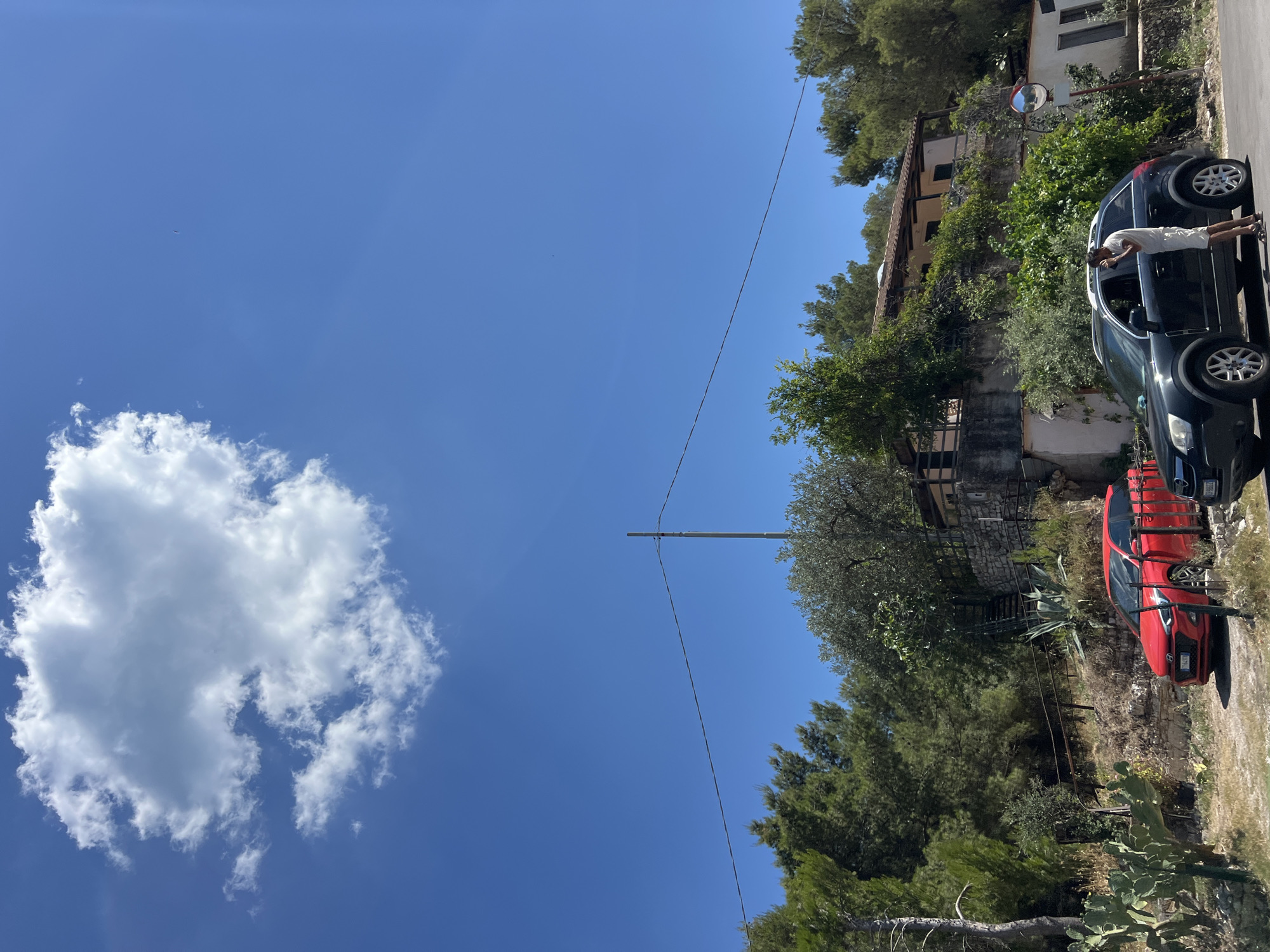This image depicts a sunny day in Mattinata, Italy, with a clear blue sky and a large, fluffy white cloud. In the foreground, two cars are parked on a dirt road: a red Hyundai i20 on the left and a dark-colored SUV on the right. A person, dressed in a white t-shirt and shorts, is standing by the driver's side door of the SUV, appearing to be on their phone.
Behind the cars, the landscape is characterized by lush greenery, with various trees and shrubs, including pine trees, olive trees, and prickly pear cacti. A stone-built house with a tiled roof is situated on a raised terrace, surrounded by vegetation. A set of outdoor stairs leads up to the house. A utility pole with a wire stretches across the frame in the mid-ground. The overall setting suggests a rural or semi-rural area within or near Mattinata, possibly on a hillside overlooking the coast, given the typical scenery of the Gargano Peninsula.