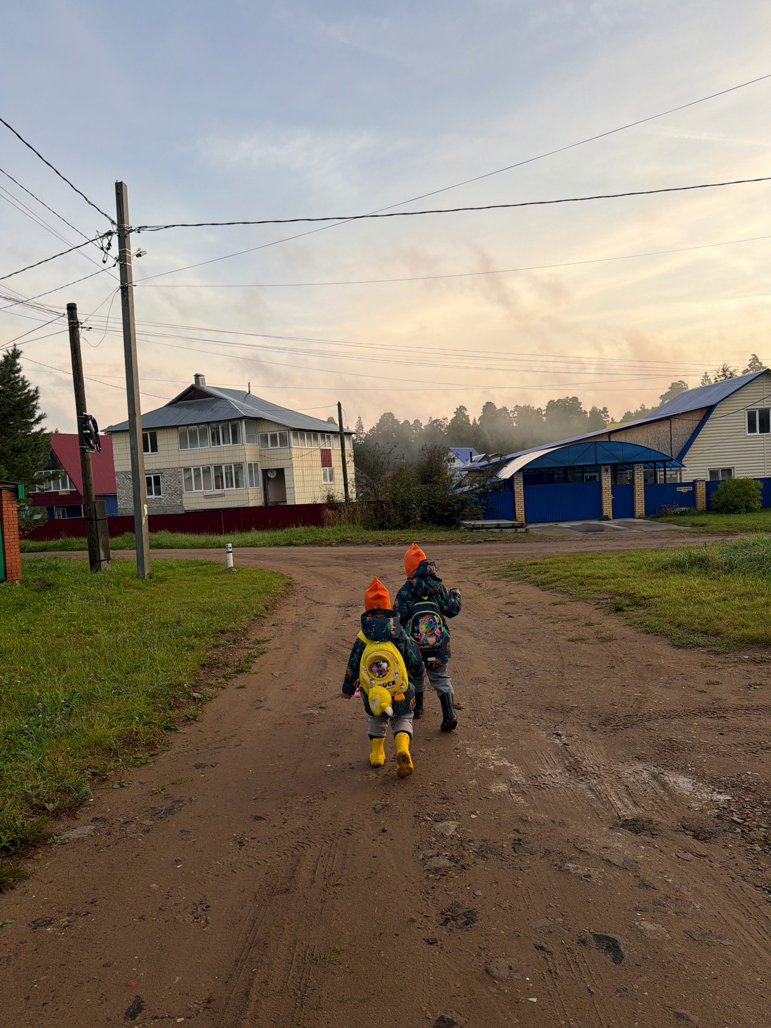 Two young children, each wearing an orange hat, a patterned jacket, and yellow boots, walk away from the camera on a dirt road. The child on the left has a yellow backpack with a duck design. The child on the right has a backpack with a dinosaur design. They are walking towards houses and trees under a hazy, overcast sky. The location is a rural area, likely on the outskirts of Chaikovsky, Russia, given the style of housing and the presence of utility poles and overhead wires. The road is uneven and muddy in places, with tire tracks visible. The houses are a mix of styles, with one larger, multi-story house in the background and smaller, more traditional homes visible. There is vegetation, including grass and trees, on either side of the road. Smoke or mist is visible in the distance over the trees.