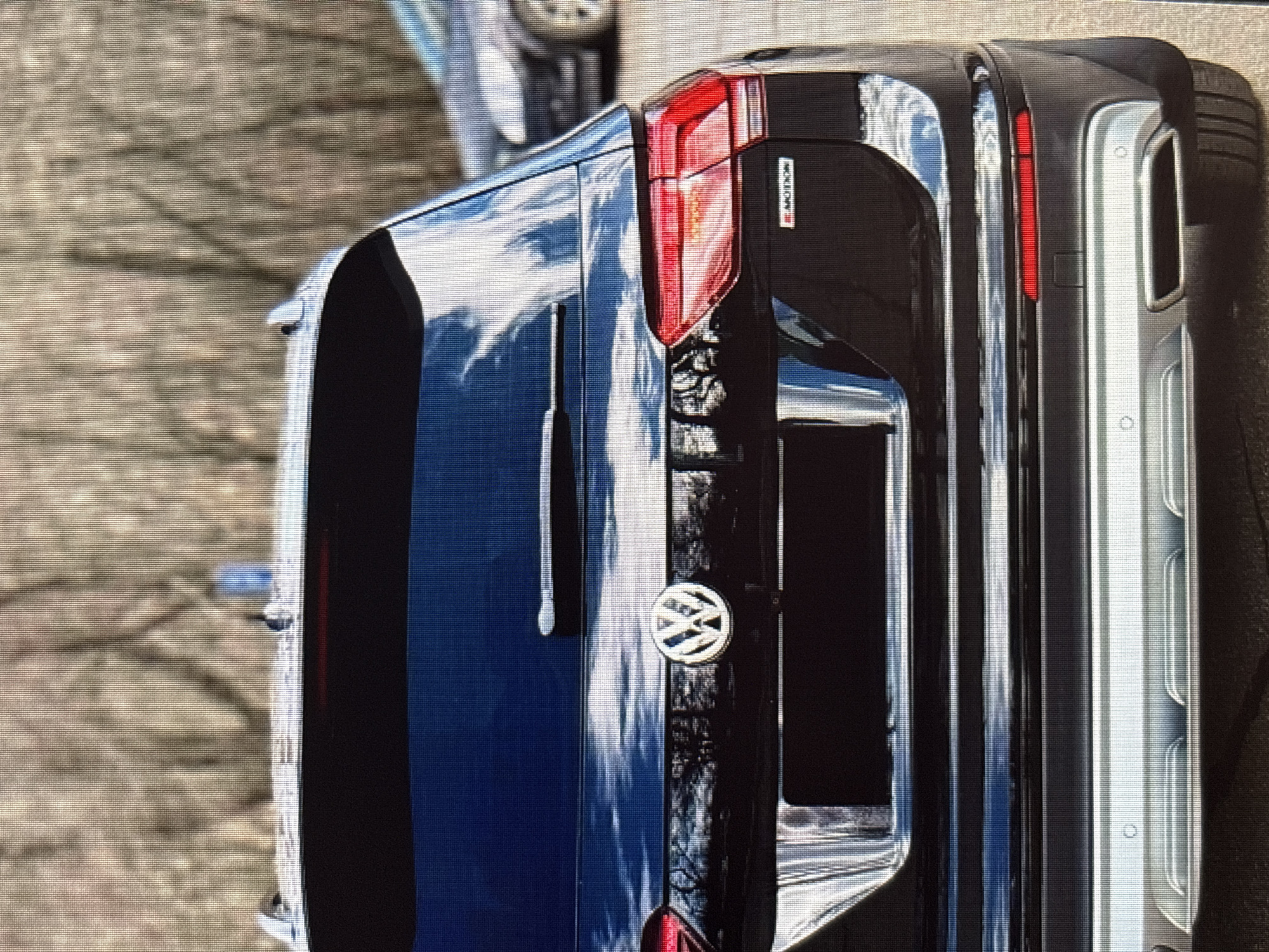 The image shows the rear view of a black Volkswagen SUV parked outdoors on a paved surface. The car has a prominent Volkswagen logo on the tailgate and a red "4MOTION" badge on the right side. The vehicle's taillights are visible, and a rear windshield wiper is present. The background consists of bare trees and foliage, suggesting a season like late autumn or winter. Another car is partially visible in the background to the right. The image lacks any discernible city landmarks, specific architectural details, or human subjects that would definitively place it in Moscow, Russia, or any other particular urban location.