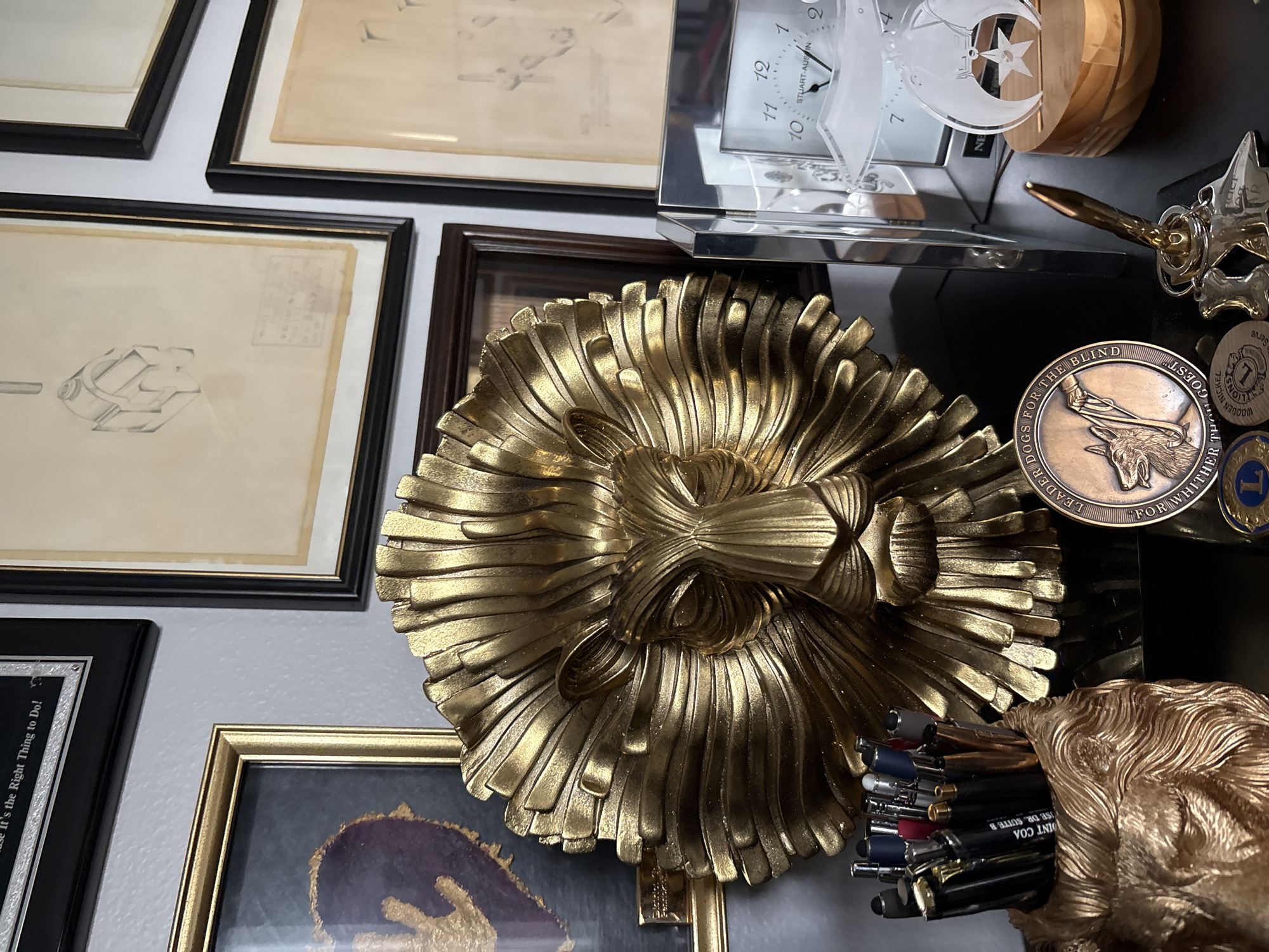 The image depicts a desk with several decorative items and office supplies, likely in a home or office setting. The primary focal point is a large, ornate gold lion head sculpture with a detailed mane. To the left of the lion head is a framed piece of art with a dark background and a gold design. In front of the lion head, a pen holder shaped like a gold lion's head is filled with various pens and writing utensils. A circular bronze medal with an image of a dog and text reading "LEADER DOGS FOR THE BLIND" is positioned in front of the pen holder. To the right of the lion head sculpture is a clear acrylic framed clock with Roman numerals. A small wooden base holds an award with a star and circle design. The background features framed artwork, including sketches. The overall impression is one of a curated display of personal or professional memorabilia. The location is identified as Sun City Center, United States of America.