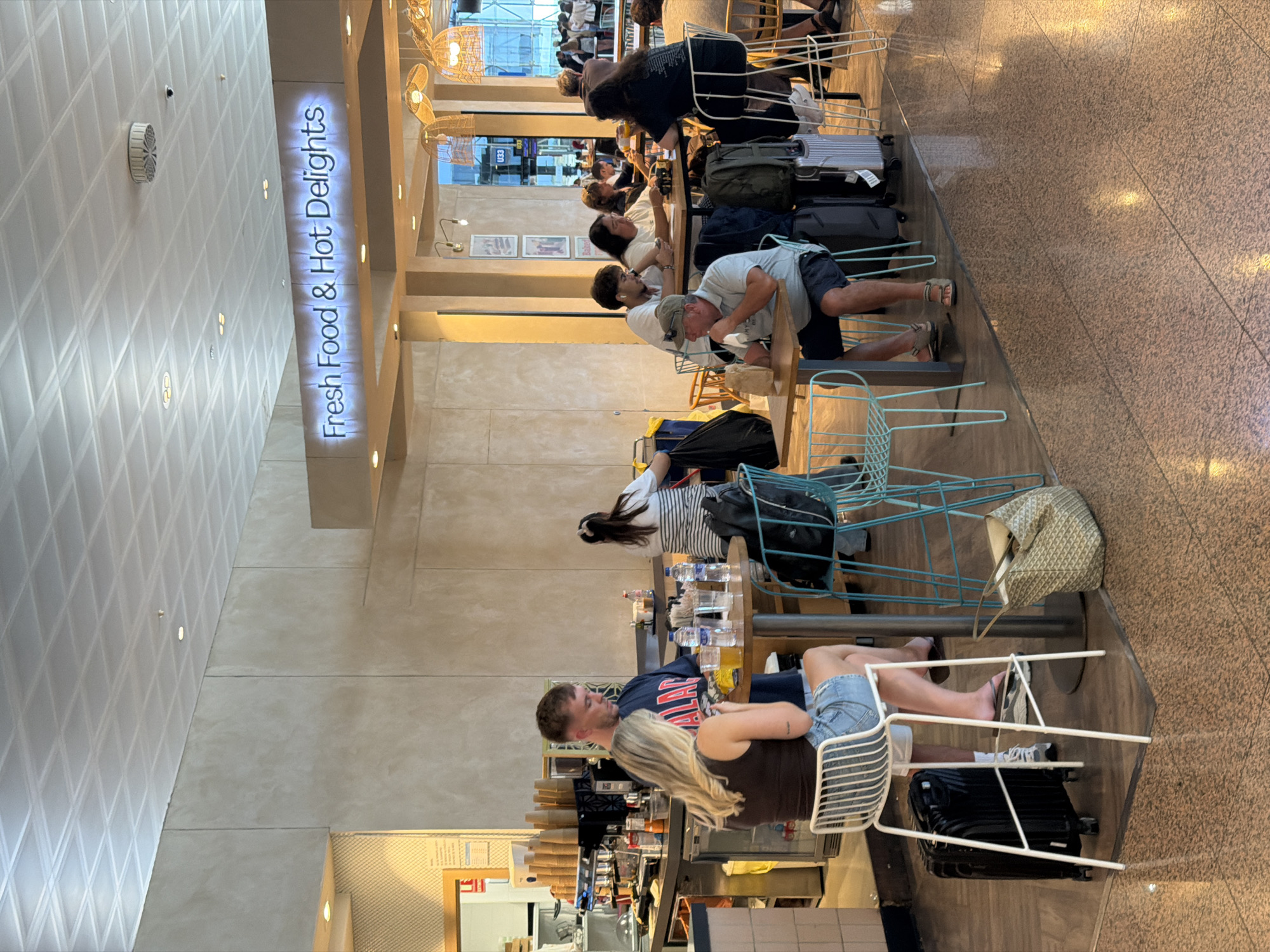 The image captures an indoor scene, likely an airport terminal in El Prat de Llobregat, Spain, given the context of travel-related elements. The setting appears to be a cafe or eatery with a "Fresh Food & Hot Delights" sign prominently displayed. Several individuals are seated at tables, engaged in various activities such as eating, drinking, and conversing. A couple is seated at a high table near the foreground, with the woman facing away from the camera and the man looking down. Other patrons are visible in the background, some at similar high tables and others at lower tables. Luggage is present, including suitcases near the tables and a large tote bag on the floor, further suggesting an airport environment. The cafe area features bar stools, tables, chairs, and what appears to be a service counter with a coffee machine and stacks of cups. The lighting is a mix of ambient overhead light and accent lighting from decorative fixtures. The floor is a polished speckled material.