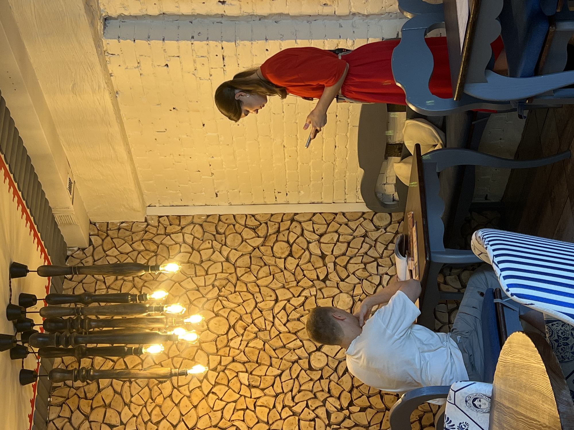 The image depicts the interior of a restaurant or cafe in Chelyabinsk, Russia. The decor features a wall covered in circular wood slices and a white brick wall. A large, dark wooden chandelier with multiple bare bulbs hangs from the ceiling. In the foreground, a man wearing a white t-shirt and light-colored pants sits at a table, his head resting on his hand, looking down at an open book. To the right, a woman in a red dress stands holding a smartphone in her hands, appearing to interact with it. She is positioned near a grey upholstered bench with cushions. Several tables and chairs are visible, with some chairs featuring striped blue and white upholstery. The lighting is warm, suggesting an indoor setting, possibly evening.