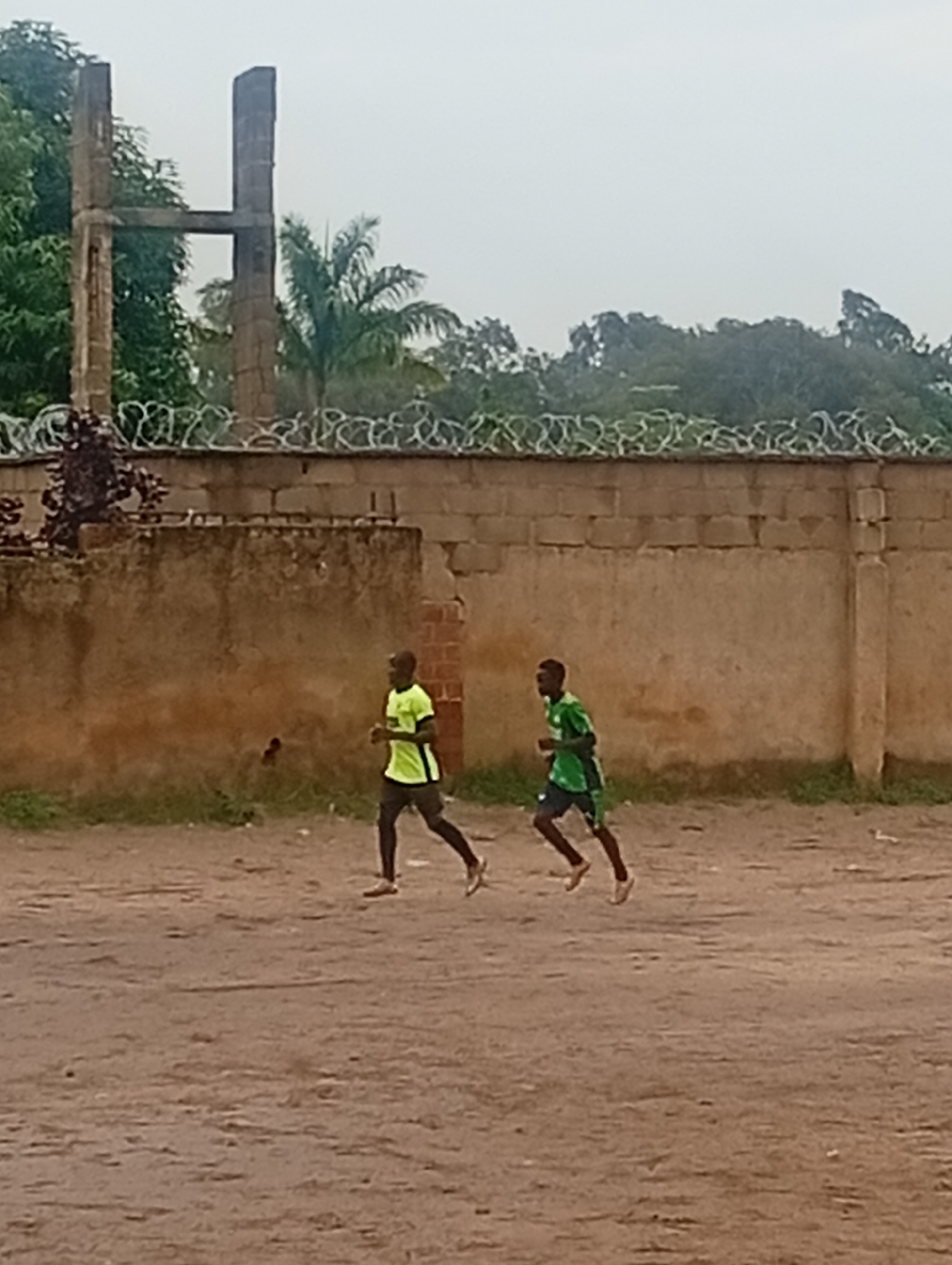 In Jos, Nigeria, two young men are captured in motion, jogging along a dusty, outdoor track. The man on the left is wearing a bright yellow t-shirt and dark shorts, his form suggesting a steady pace. To his right, a second man, clad in a green and black t-shirt and shorts, runs with a slightly more dynamic stride. They are positioned in front of a weathered, beige brick wall topped with a band of barbed wire, creating a stark contrast with the lush green foliage and palm trees visible behind it. In the background, partially obscured by trees, stands a tall, skeletal structure resembling a cross, possibly a remnant of a past monument. The overall environment suggests an overcast day in a somewhat undeveloped or utilitarian area of the city.