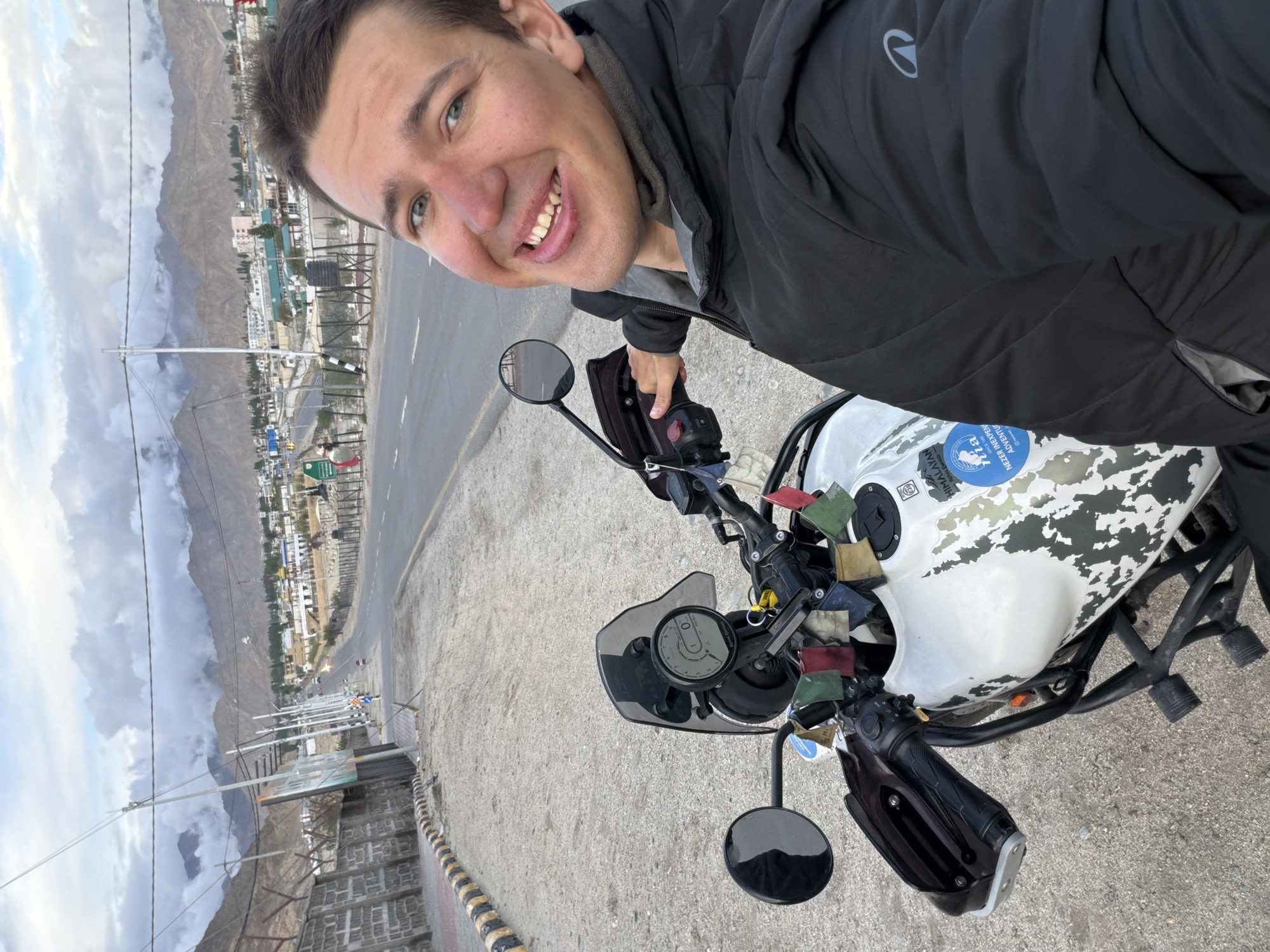 A young Caucasian man with blue eyes and fair skin is taking a selfie while sitting on a white Royal Enfield Himalayan motorcycle. He is wearing a black puffer jacket. His mouth is open, revealing teeth, and he appears to be smiling. The motorcycle has a black windscreen, a digital display showing "0", and prayer flags attached to the handlebars. The bike is parked on a gravel surface next to a paved road. In the background, a town with buildings and mountains can be seen under a cloudy sky. The scene is located on a road leading into the town of Spituk, India, as indicated by a signpost with "Spituk" and directional arrows. The road is lined with a stone wall and metal poles supporting overhead wires.