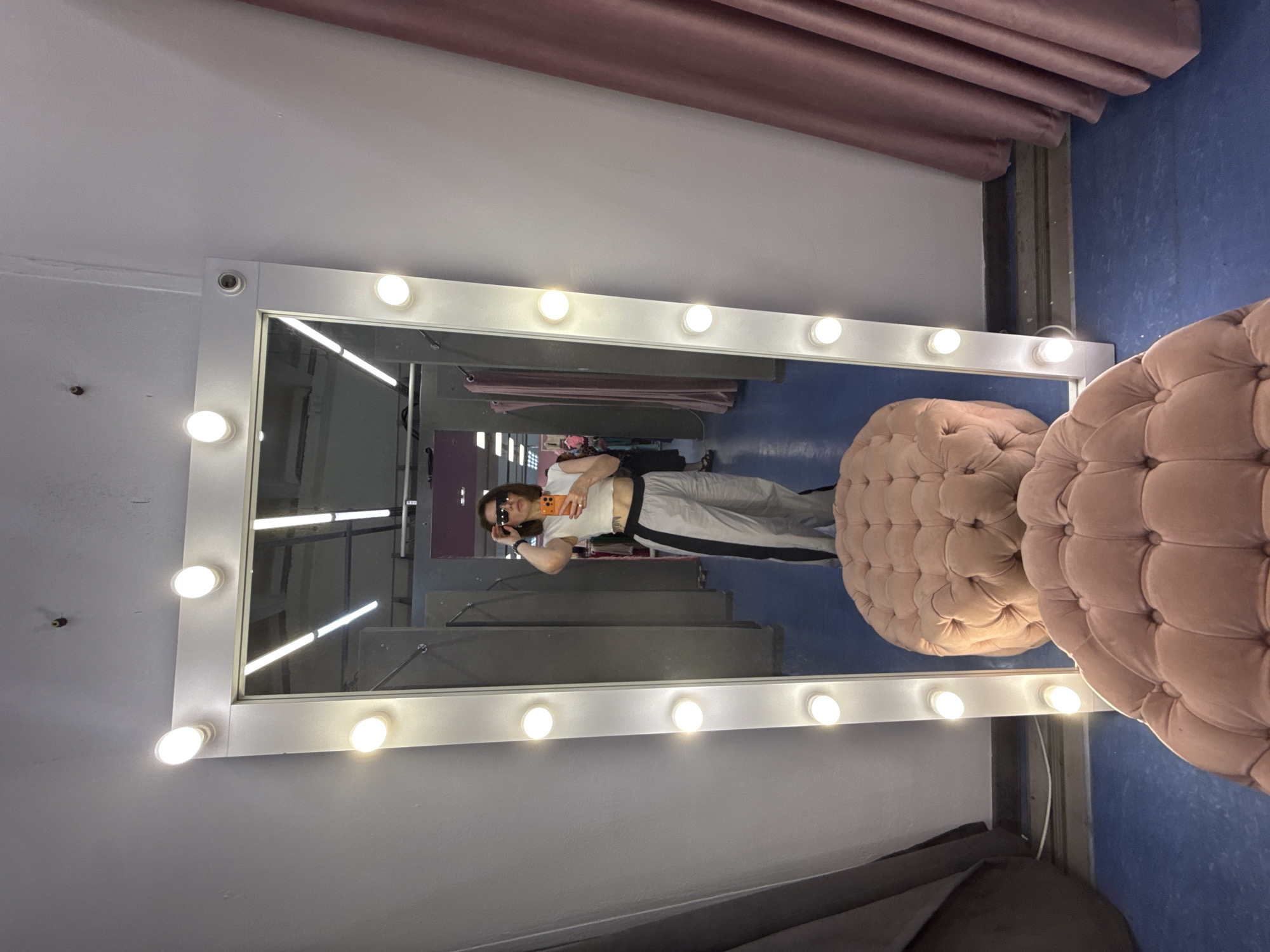 The image shows a woman reflected in a large mirror with lights around its perimeter. The woman is taking a selfie with her phone. She is wearing sunglasses, a white top, and gray sweatpants with black stripes. The environment appears to be a dressing room or retail space. Behind the woman in the reflection, there is a glimpse of a retail corridor with hanging clothes. Next to the mirror are two round, tufted ottomans in a muted pink color. Above the mirror, there is a draped piece of material, also in a muted pink tone. The background color to the right of the mirror is a dark blue. The image was taken in Izhevsk, Russia.