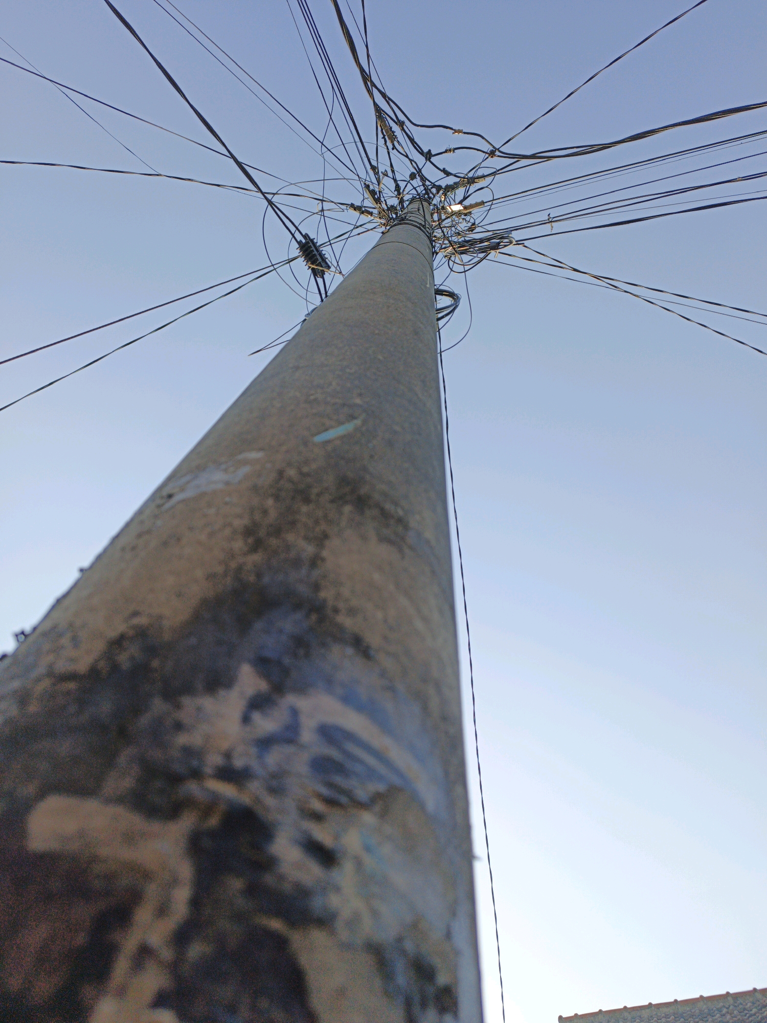 A low-angle, close-up shot captures a utility pole dominating the frame, its surface weathered and marked with patches of peeling paint and grime. The pole extends upwards, disappearing into a chaotic tangle of black electrical wires against a clear, pale blue sky. The wires, numerous and haphazardly arranged, fan out in various directions, suggesting a complex and possibly unorganized distribution network. The foreground of the pole is out of focus, with a significant portion of its textured and discolored surface filling the left side of the image. In the far lower right corner, a sliver of a tiled roof is visible, hinting at the presence of a building or structure below. The scene appears to be an outdoor urban environment, possibly in Ketegan, Indonesia, given the context.