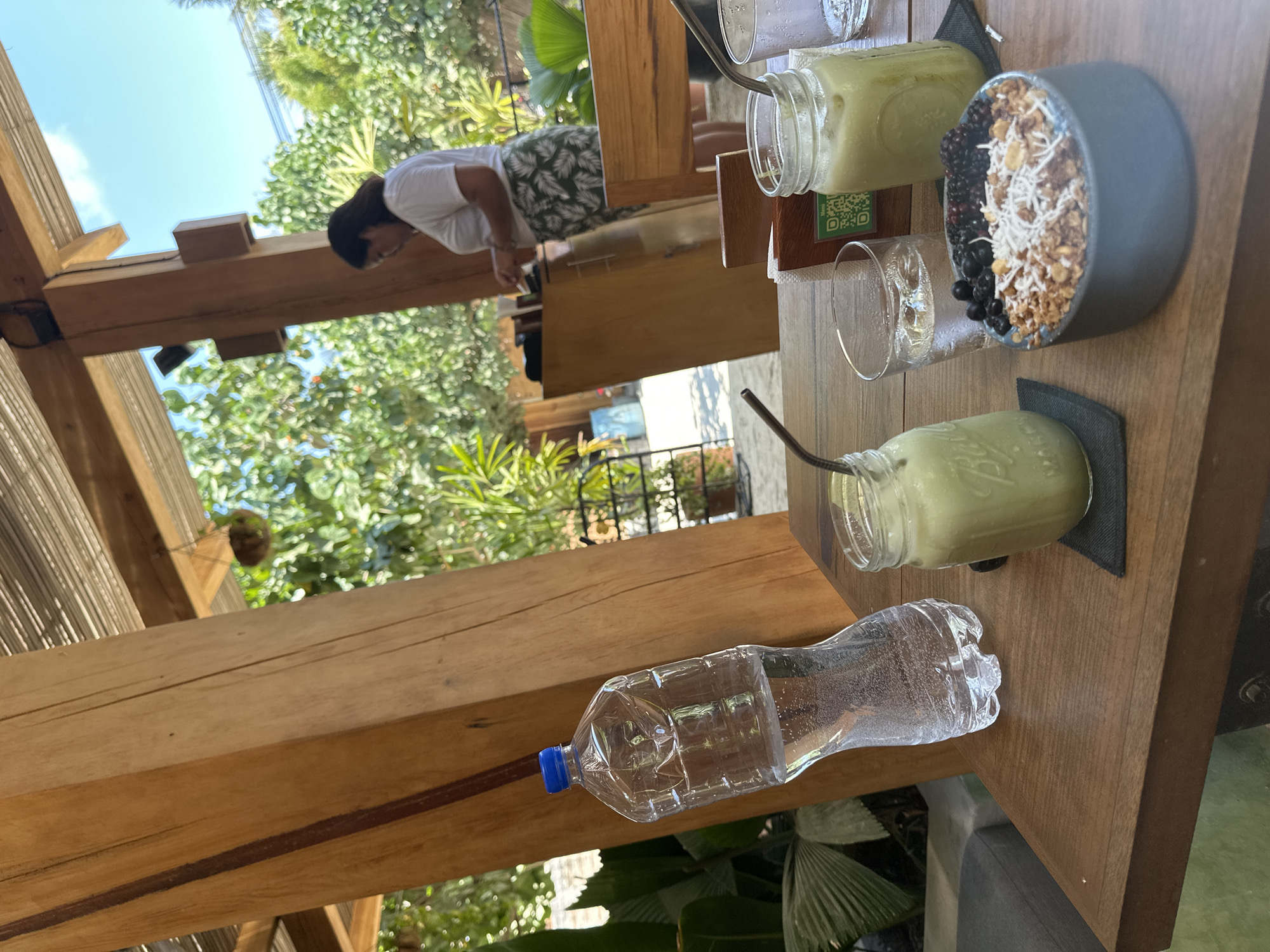 The image shows a wooden table with a bottle of water, two green smoothie-like drinks in jars with straws, and a bowl of what appears to be granola, coconut flakes, and blackberries on top. A woman in a white shirt and patterned shorts stands beyond the table. The setting seems to be outdoors, with trees and sky visible in the background. The location is Puerto Escondido, Mexico.