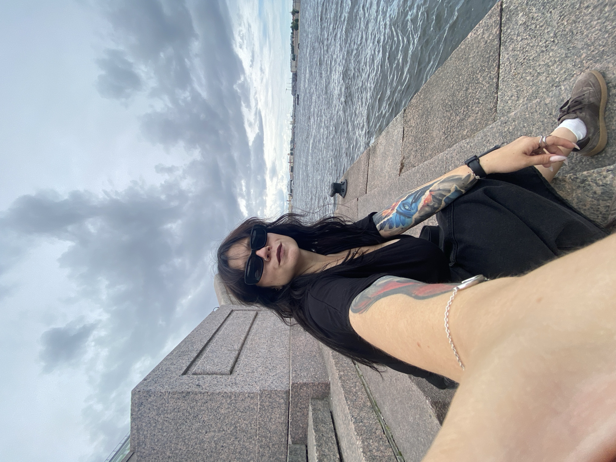 A woman with long dark hair, wearing black sunglasses, a black t-shirt, and black pants, sits on the granite steps of a waterfront embankment in Saint Petersburg, Russia. She has tattoos on her arms, including one on her left arm with a blue bird motif. She is wearing a silver bracelet on her left wrist and rings on her fingers. Her right foot is visible, wearing a brown shoe and white socks. The sea is to her right, with a city skyline in the distance. The sky is overcast with dark clouds. The steps are made of rough, pinkish-grey granite.