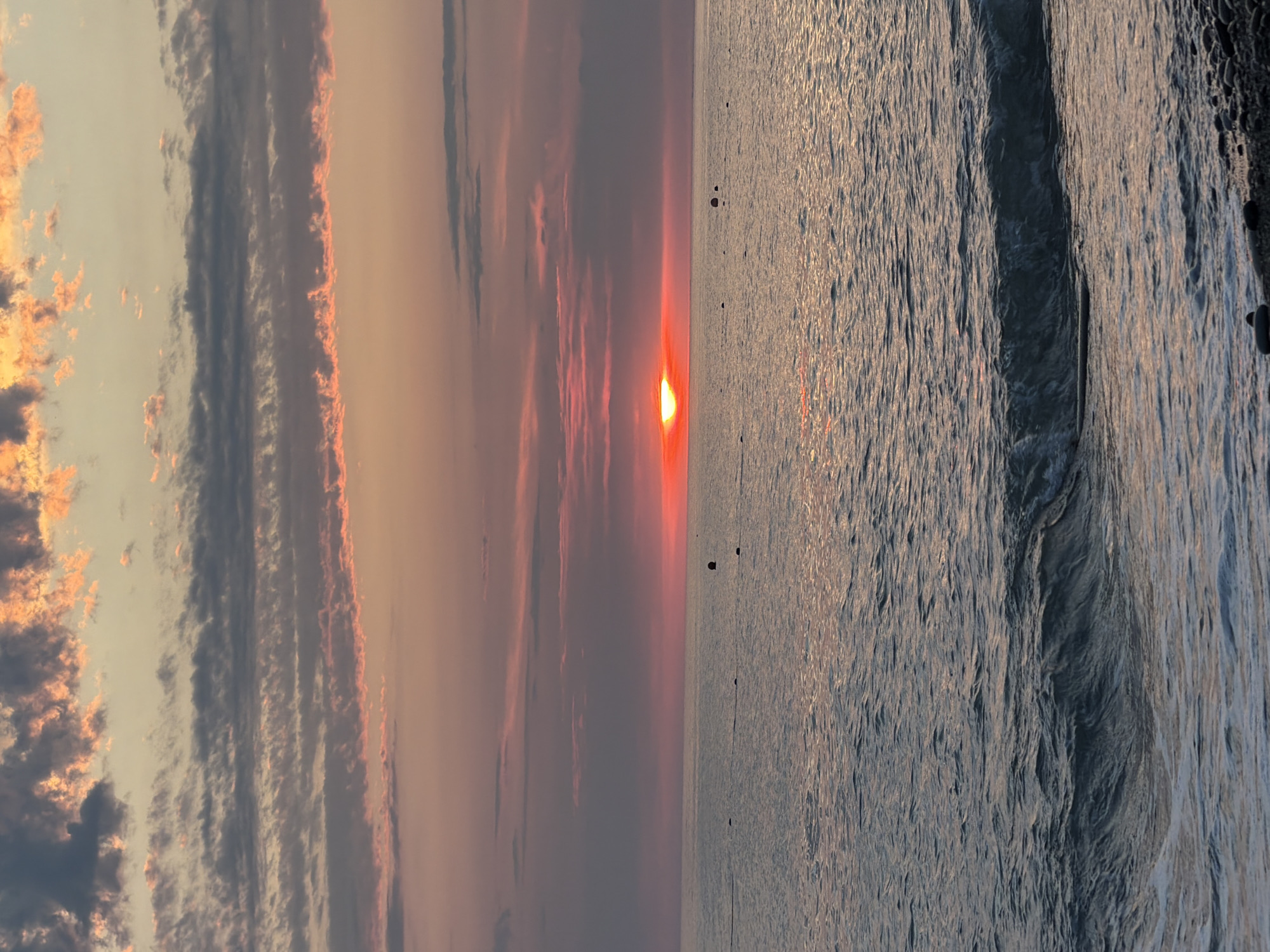 A sunset over the Black Sea in Sochi, Russia, is captured from a pebbly shore. The sun, a vibrant red orb, is partially obscured by horizontal clouds, casting a warm glow across the sky. The water is choppy with a gentle wave breaking near the shore. Several buoys are visible on the surface of the water. The sky is filled with streaks of pink, orange, and grey clouds, indicating a dramatic end to the day. The scene is tranquil and serene, evoking a sense of peace and natural beauty.