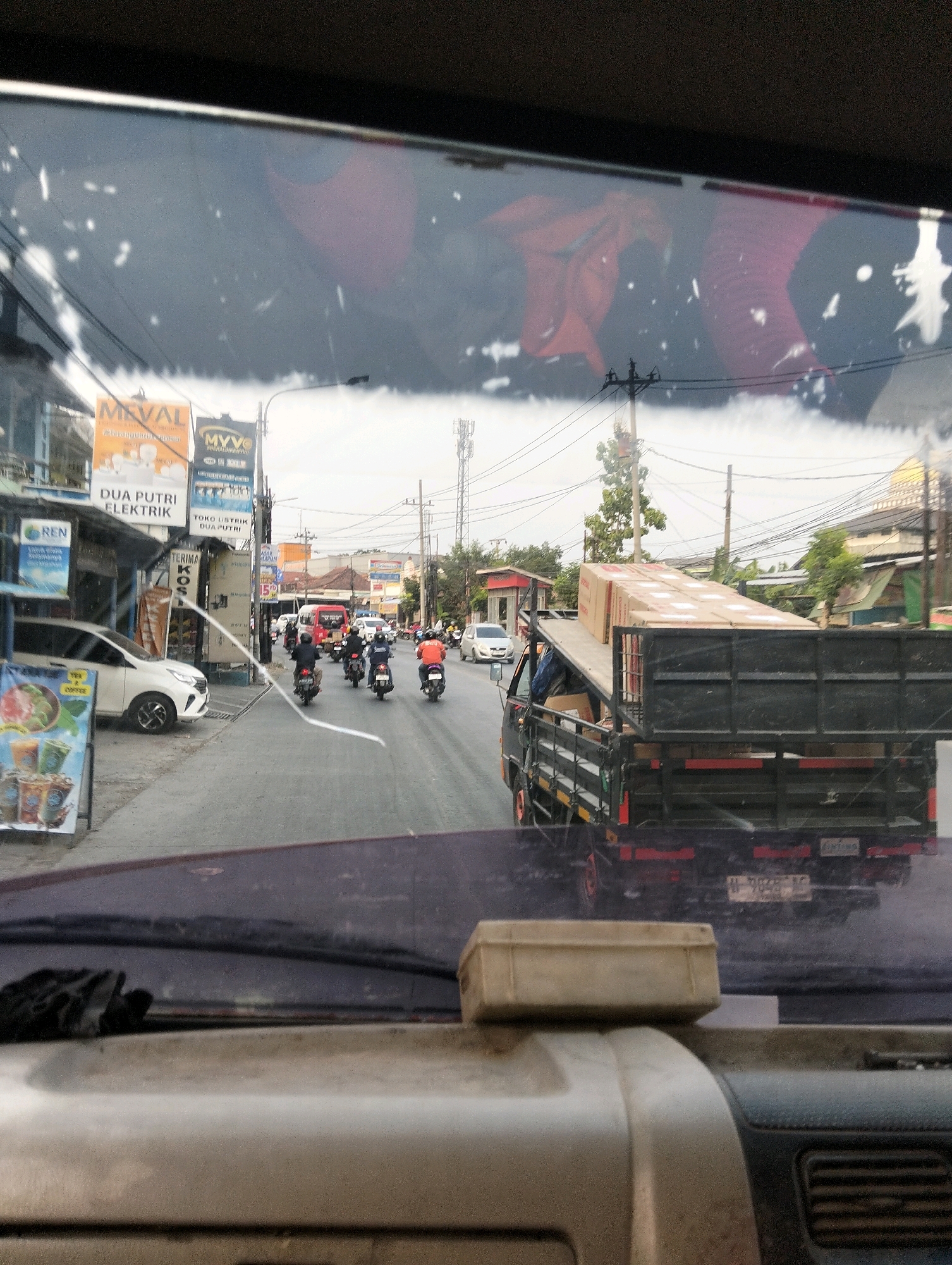 This image, captured from inside a vehicle, depicts a street scene in Semarang, Indonesia. The foreground shows the dashboard and windshield of the vehicle, with reflections of objects, possibly the interior of the car and a red draped object, visible on the windshield. Outside the vehicle, a road is visible with several motorcycles carrying riders and a truck loaded with boxes. On the left side of the road, there are shops and a white car parked. Billboards with text, some in Indonesian, are also present. The environment appears to be a busy urban street with buildings and overhead power lines. The sky is overcast, suggesting a cloudy day.