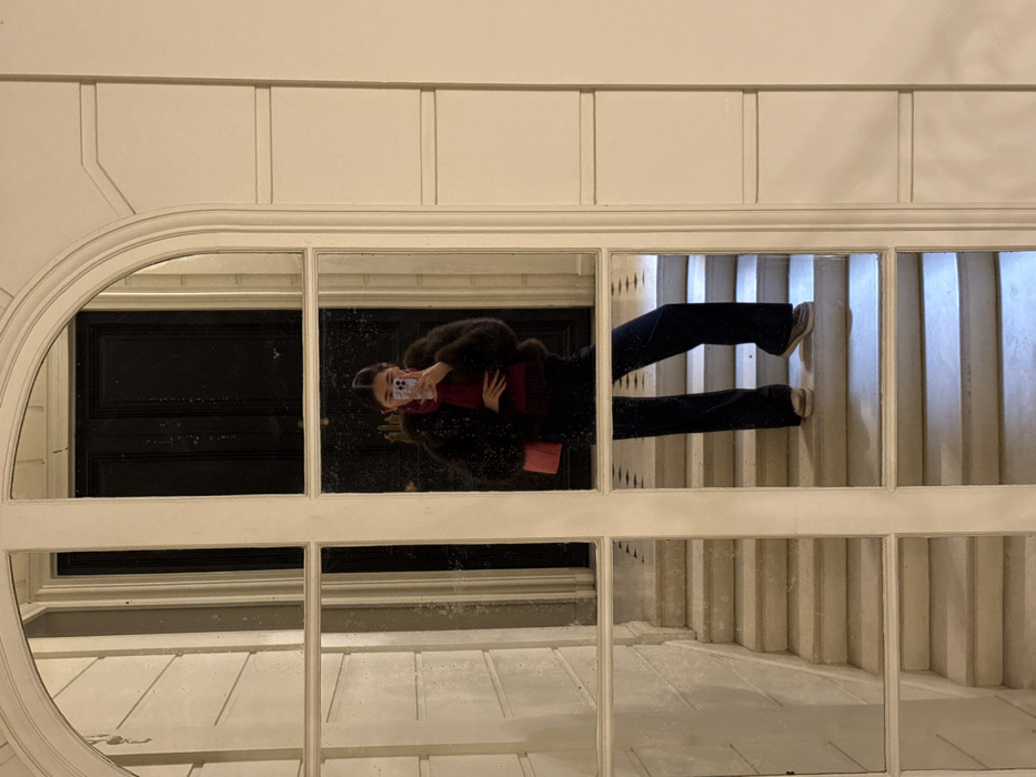 A person is reflected in an arched, multi-pane window or mirror, taking a self-portrait with a light-colored smartphone. The setting is an interior entrance hall or hallway, identified by Haussmannian architectural elements typical of Paris, France. The individual wears a dark brown fur-textured jacket, a deep red top, dark blue wide-leg trousers, and light-toned flat shoes, accessorized with a small pink shoulder bag. Their dark hair is styled back. The reflection reveals a dark paneled double door in the background and light-colored stone walls adorned with classical moldings. A light-toned stone staircase is visible ascending to the left and beneath the person's feet. The mirror's panes are separated by thick white mullions and transoms, and the glass appears aged.
