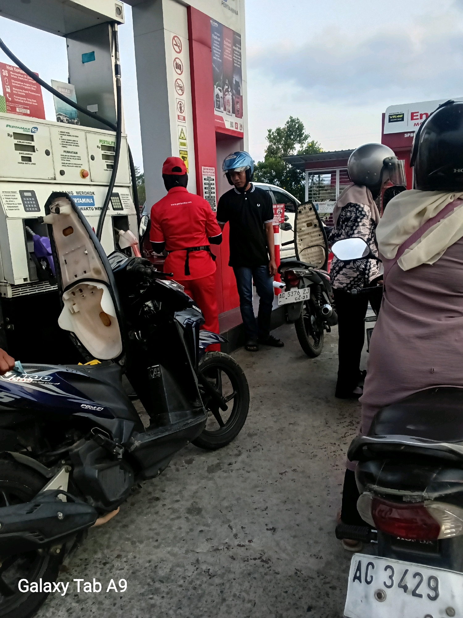 The image depicts a scene at a Pertamina gas station in Trenggalek, Indonesia, during daylight hours with an overcast sky. Two motorcycles are present; one is in the foreground with its seat open, and another is further back. A gas pump, labeled "PERTAMINA GREEN 95" and "PERTALITE", is visible with fuel hoses.
There are at least four individuals in the scene. One person, wearing a red uniform with "www.pertamina.com" printed on it, is standing near the gas pump, appearing to be a gas station attendant. Another person, wearing a black polo shirt and blue helmet, is standing behind the first motorcycle. Two other individuals are on or near the second motorcycle; one is wearing a headscarf and a patterned top, and the other is partially visible in a light-colored head covering. One of the motorcycles has a license plate reading "AG 5376 ZJ" and "02-31", and the other has "AC 3429". The ground is paved with concrete.