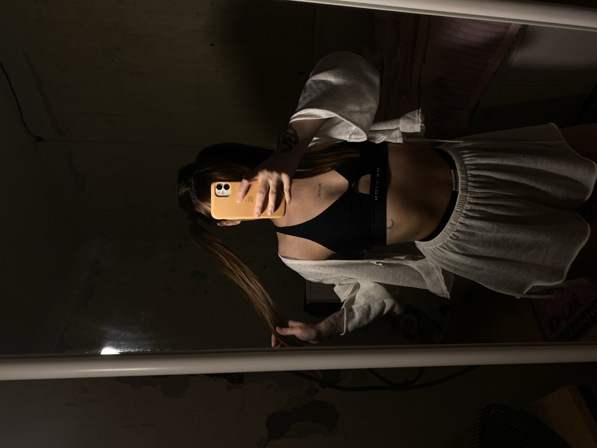 A person with long hair and a light-skinned complexion is visible in a mirror selfie. The individual is wearing a black sports bra and light-colored shorts, with a white shirt draped over their shoulders. They are holding a yellow smartphone in front of their face, obscuring their features. The background is dimly lit and reflects a cluttered room. The location is unknown and cannot be determined from the image. The environment consists of items in the background and mirror frame.