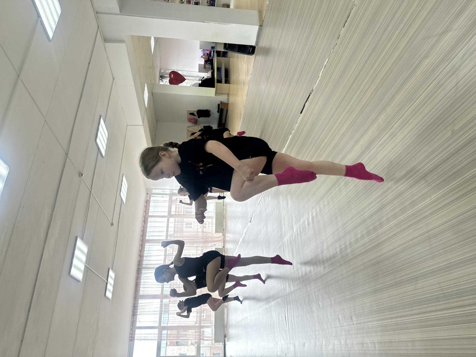 The image captures an indoor scene of several young girls practicing dance or rhythmic gymnastics in a studio in Votkinsk, Russia. The photograph is oriented vertically, rotated 90 degrees counter-clockwise, giving the impression of the subjects hovering above the floor.
The main subjects are multiple pre-teen girls, all dressed in black athletic attire, consisting of t-shirts or leotards and shorts. Most of them are wearing bright pink ankle socks, though some have black socks. They are engaged in a synchronized pose, bent at the waist with one leg extended upwards, demonstrating flexibility and balance. Their faces show focus and concentration on their movements.
The setting is a well-lit studio featuring a light-colored, possibly wooden or laminate floor with a striped pattern. The walls are white, and large rectangular windows along one side allow ample natural light into the room. Additional illumination comes from rectangular fluorescent light fixtures on the ceiling. A long mirror on one wall reflects the girls and the studio space. In the background, near some shelves, a red heart-shaped balloon and a mobile phone are visible. The time of day appears to be daytime due to the natural light. There is no visible text in the image.