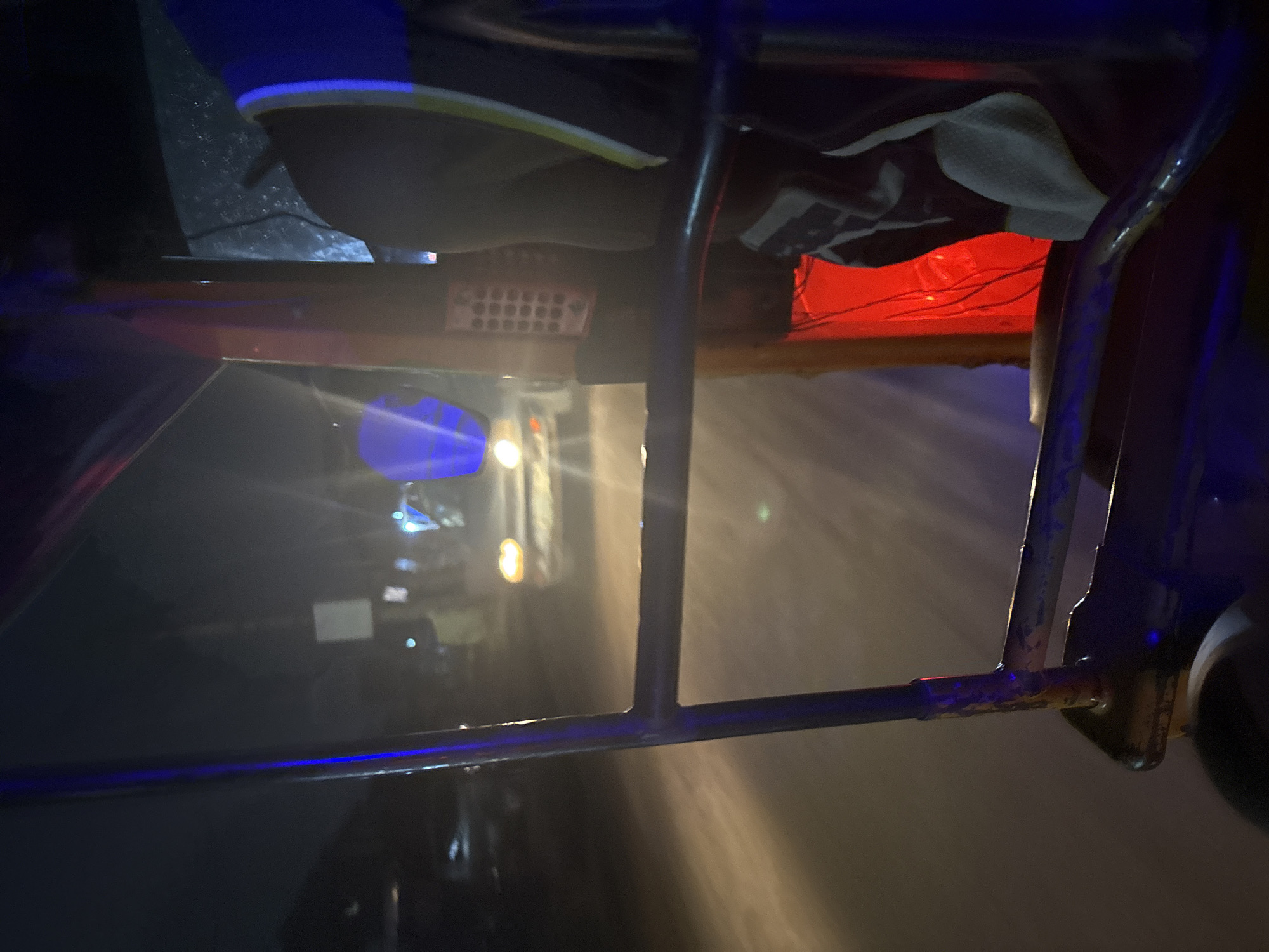 The image depicts a nighttime scene from within a vehicle, likely a tuk-tuk, looking out onto a dimly lit street in Nkpor, Nigeria. The foreground is dominated by the interior of the vehicle, with metal bars and a blue light strip visible. The street ahead is blurred, suggesting motion, and is illuminated by the headlights of an oncoming vehicle and ambient streetlights. The overall atmosphere is hazy and somewhat ethereal due to the low light conditions and possibly mist or dust. The environment appears to be an urban or semi-urban setting with buildings visible in the background, but they are indistinct. The scene is captured with a shallow depth of field, emphasizing the interior of the vehicle and the blurry road.