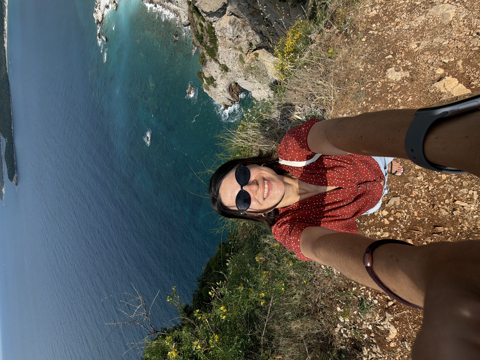 This high-angle selfie captures a smiling woman from a clifftop overlooking the Adriatic Sea near Budva, Montenegro. The woman, with dark hair and sunglasses, looks up at the camera with a joyful expression. She is wearing a reddish-brown top with white polka dots and what appear to be light-colored shorts or a skirt, along with flip-flops on her visible right foot. A grey-banded smartwatch is on her left wrist, and a dark brown hair tie or bracelet is on her right.
The perspective showcases a dramatic coastal landscape. Directly below, a steep, rocky cliff face drops sharply to the sea, where turquoise waters meet the shore with gentle waves. Further out, the water transitions to a deep, serene blue. Scattered rocks and small islands are visible along the coastline. On the left side of the frame, a distant, greener landmass or island stretches across the horizon.
The ground where the woman stands is dry, rocky, and covered with sparse, hardy vegetation including dry grasses and small bushes with yellow and purple flowers, characteristic of a Mediterranean climate. The bright sunlight suggests a clear, sunny day, likely during late morning or early afternoon. The image conveys a sense of adventure and appreciation for scenic natural beauty. There is no visible text in the image.