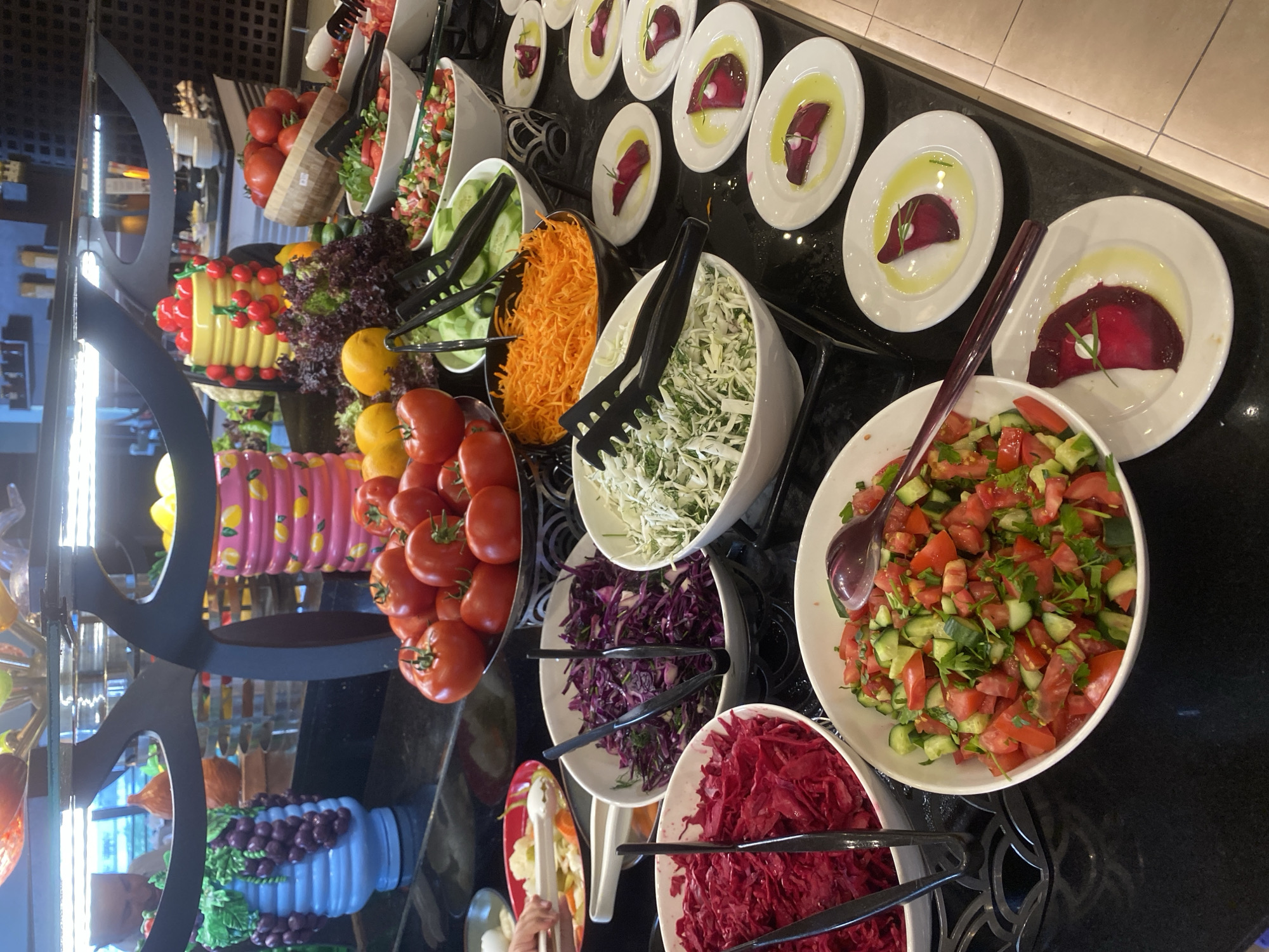 The image shows a buffet-style food service, likely in a restaurant or hotel in Aksu, Turkey. The display features various salads and fresh produce. In the foreground, there are several white bowls containing chopped tomatoes, cucumbers, and herbs. To the left, a large bowl of shredded red cabbage is visible, with black tongs resting inside. Next to it, another bowl holds a similar red shredded vegetable, possibly pickled cabbage or beetroot. Further back, a bowl of shredded carrots is prominent. In the background, a display of whole tomatoes in a decorative bowl can be seen. Several small white plates, each holding a slice of beetroot drizzled with oil and garnished with a sprig of herb, are arranged in a line. Decorative elements, including a large blue vase with artificial grapes and colorful ceramic pots, add to the setting. A person's hand is visible in the upper left corner, reaching for food from a serving dish. The environment appears to be indoors with bright overhead lighting.
