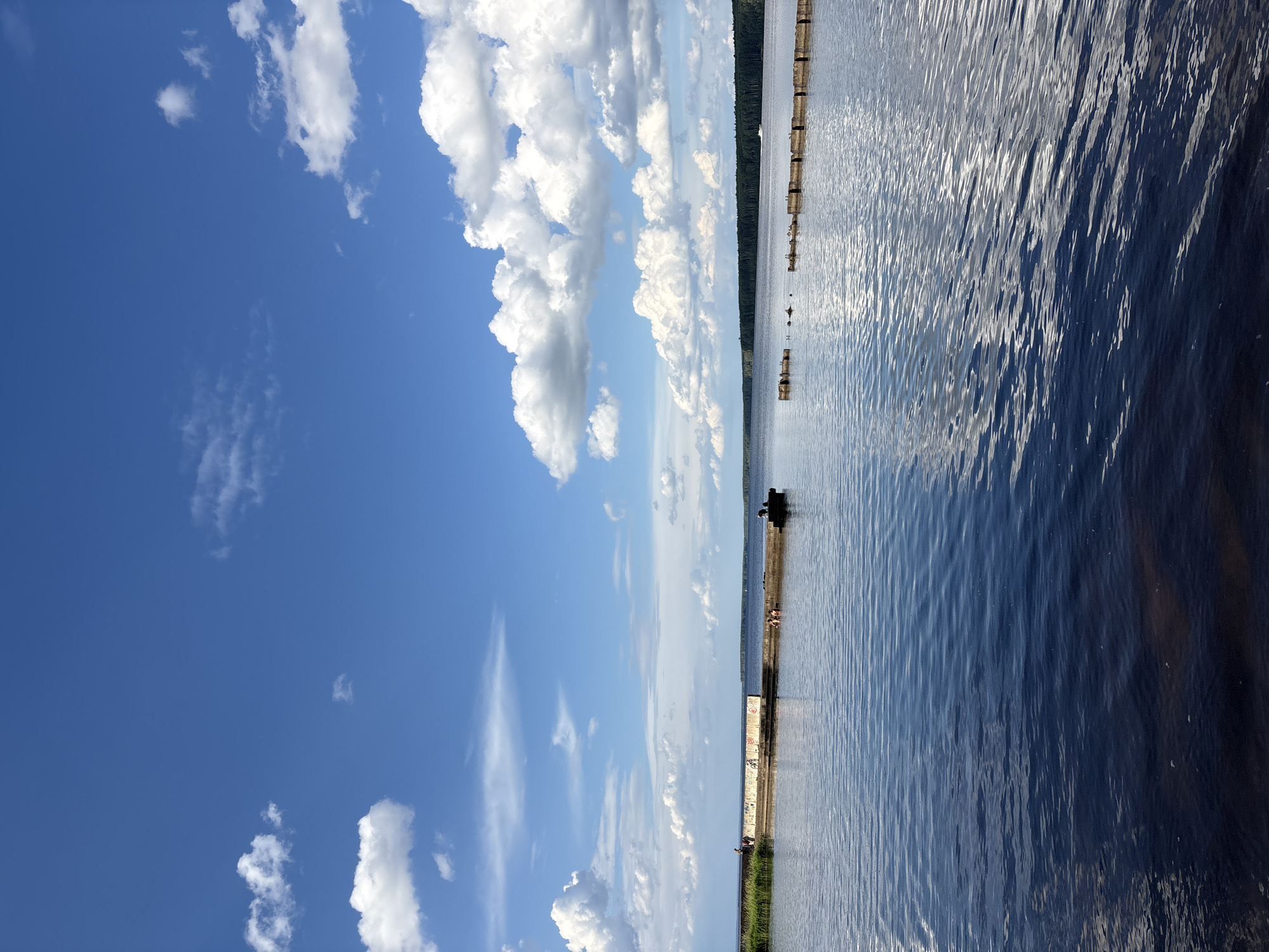 The image depicts a wide expanse of dark blue water under a bright blue sky dotted with fluffy white clouds. In the distance, a dense line of dark green trees forms the horizon.
In the foreground, the water is calm with gentle ripples, reflecting the light from the sky. A submerged structure, possibly part of a pier or breakwater, is visible in the mid-ground, extending from the right side of the frame towards the center. This structure appears to be made of concrete segments and has several birds resting on it.
Further to the left, a longer, concrete pier juts out into the water. Several people are visible on this pier. One person stands near the far end on the left, and a small group appears to be sitting or wading in the water near the center of the pier. Another person is seated at the right end of this pier. There is also a bicycle propped up against the side of this pier.
The overall scene suggests a tranquil summer day at a waterfront location, possibly a lake or river, in Chaikovsky, Russia. The presence of a pier and people enjoying the water indicates a recreational area.