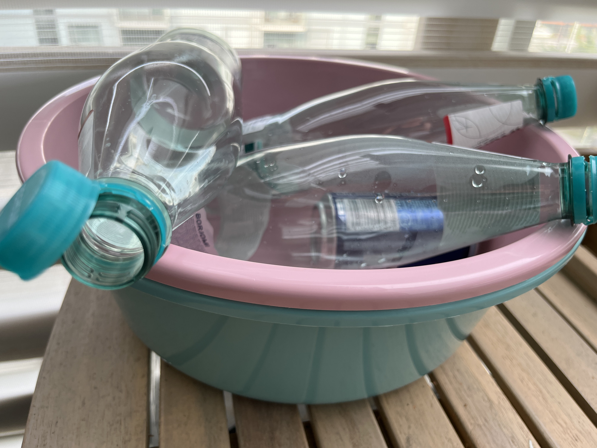 Two empty plastic water bottles, each with a turquoise cap, are placed inside a pink and teal plastic basin. The basin sits on a wooden surface, likely a table, which is partially visible in the foreground and background. The bottles are clear and contain some residual water droplets, suggesting they may have been recently emptied or rinsed. One bottle is positioned diagonally across the basin, with its opening facing the viewer. The other bottle lies beneath it, also within the basin. The background shows a blurred view of blinds and windows, indicating an indoor setting. There are no people or animals present in the image. The overall scene appears to be domestic, possibly in a kitchen or bathroom area, given the presence of the basin and water bottles. The location is not specified as Helsinki, Finland, as this information is not derivable from the visual content of the image.