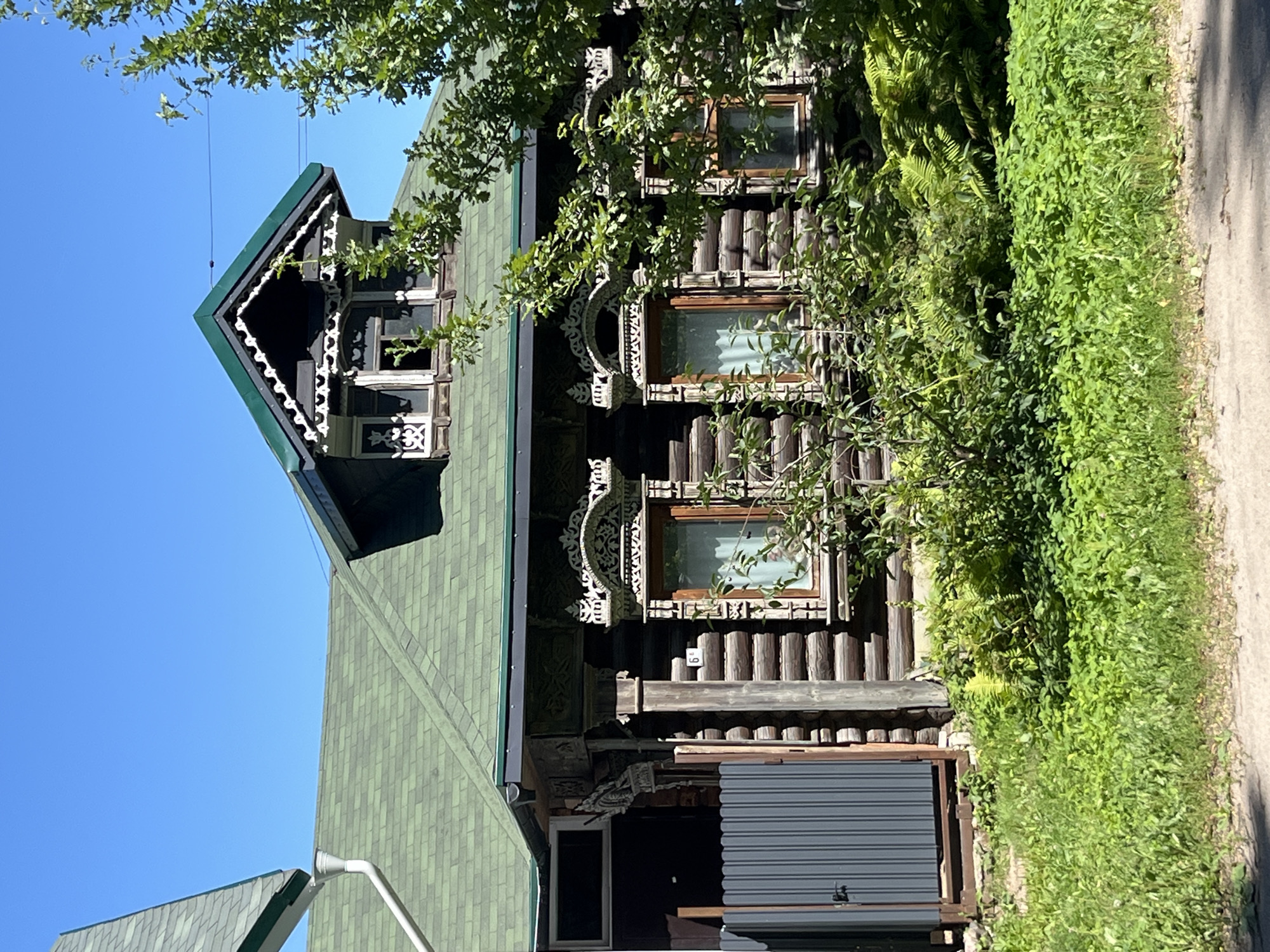 The image shows a traditional wooden house with intricate carvings around the windows and under the eaves. The house has a green shingled roof and a dark-colored dormer window protruding from the upper story. A modern gray metal gate stands to the left of the house, partially obscuring a darker section of the building. Lush green foliage, including ferns and leafy bushes, surrounds the base of the house and extends into the foreground. The sky above is a clear, bright blue, and a large tree with green leaves is visible on the right side of the frame, partially obscuring the house. The scene appears to be a sunny day in a rural or semi-rural setting, possibly in Ново-Никольское, Russia, given the architectural style. There are no people visible in the image.