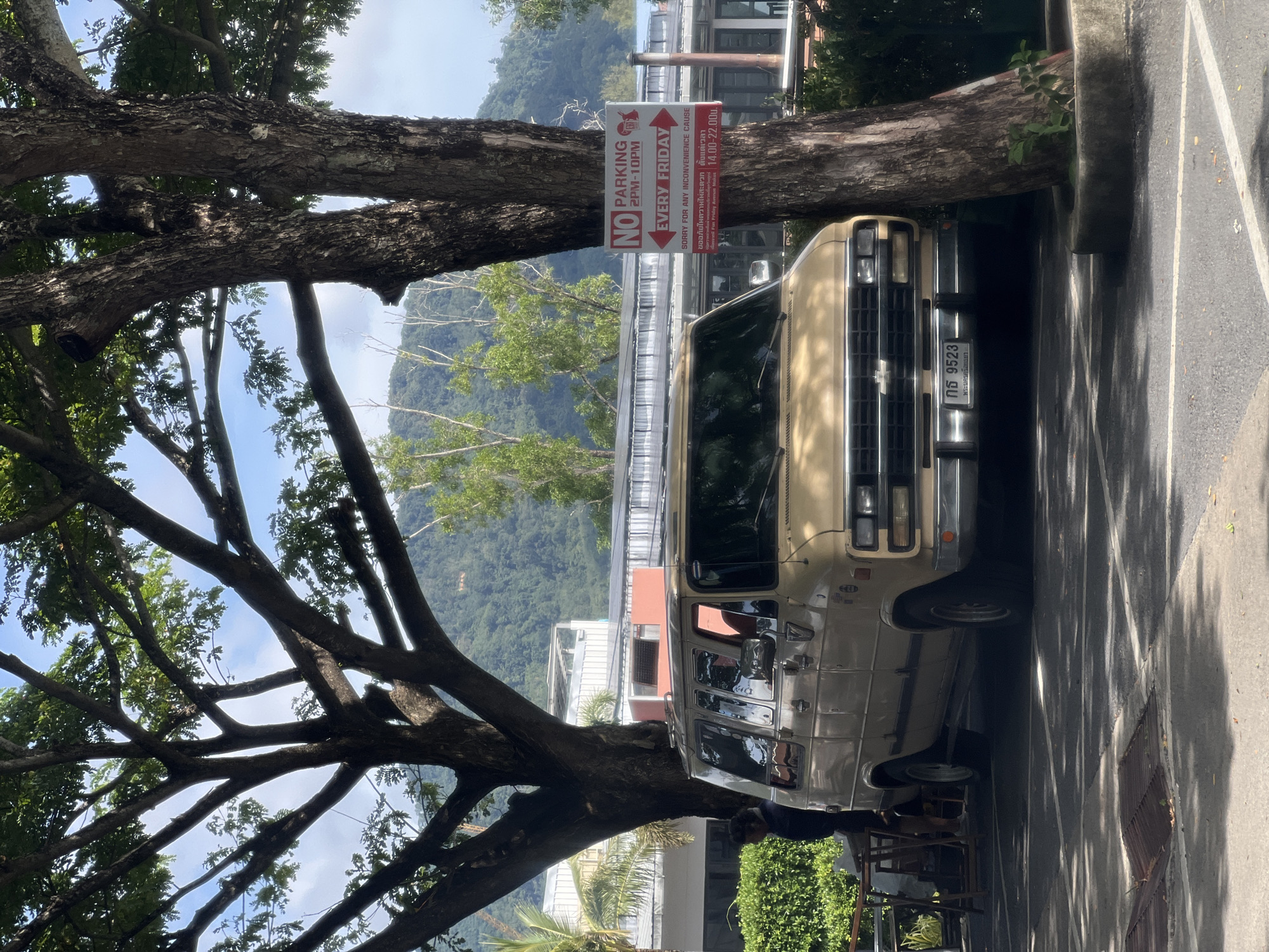 A tan van is parked under trees. A sign on one of the trees reads "NO PARKING 2PM-10PM EVERY FRIDAY". The van has a license plate with the numbers "กอ 9523". There is a person standing near the van, partially obscured by a table and chairs. In the background, there are buildings and lush green hills. The ground is asphalt with white parking lines. Sunlight filters through the leaves of the trees, casting dappled shadows.
