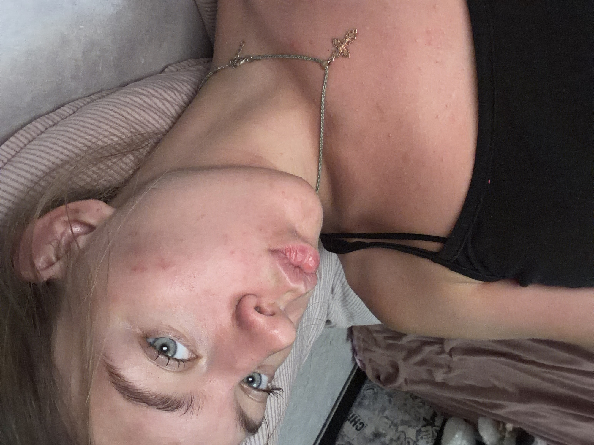 The image is a close-up, low-angle selfie of a young woman lying down. She has fair skin with some redness and blemishes, light brown hair, and striking blue eyes. She is wearing a black tank top and a delicate silver necklace with a cross pendant. Her lips are slightly parted in a pout. The background appears to be a bed with striped bedding and a glimpse of patterned fabric. The image does not contain any information about the location in Rostov-on-Don, Russia.