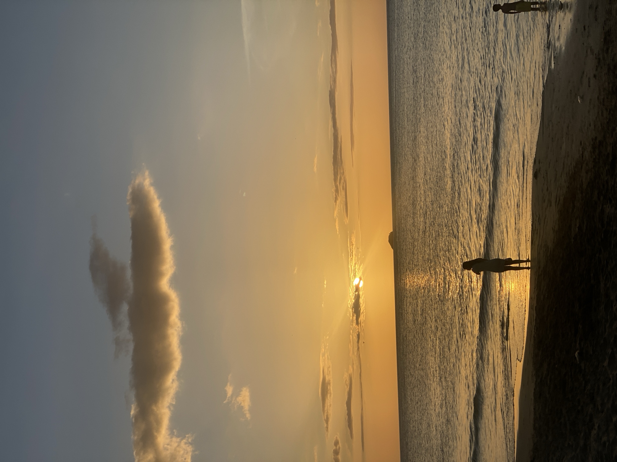 A wide shot captures a tranquil sunset over the ocean at Pedro González, Venezuela. The sun, partially obscured by clouds, casts a warm golden glow across the sky and water, with visible sun rays fanning out. A distant, small island is visible on the horizon. In the foreground, two individuals are present on the sandy beach. A woman stands near the water's edge, facing the ocean, with a small bag at her side. To the right, a child stands partially submerged in the shallow water, looking towards the ocean. The waves are gentle, lapping onto the dark sand. The overall scene is serene and evokes a sense of peace as day transitions to evening.