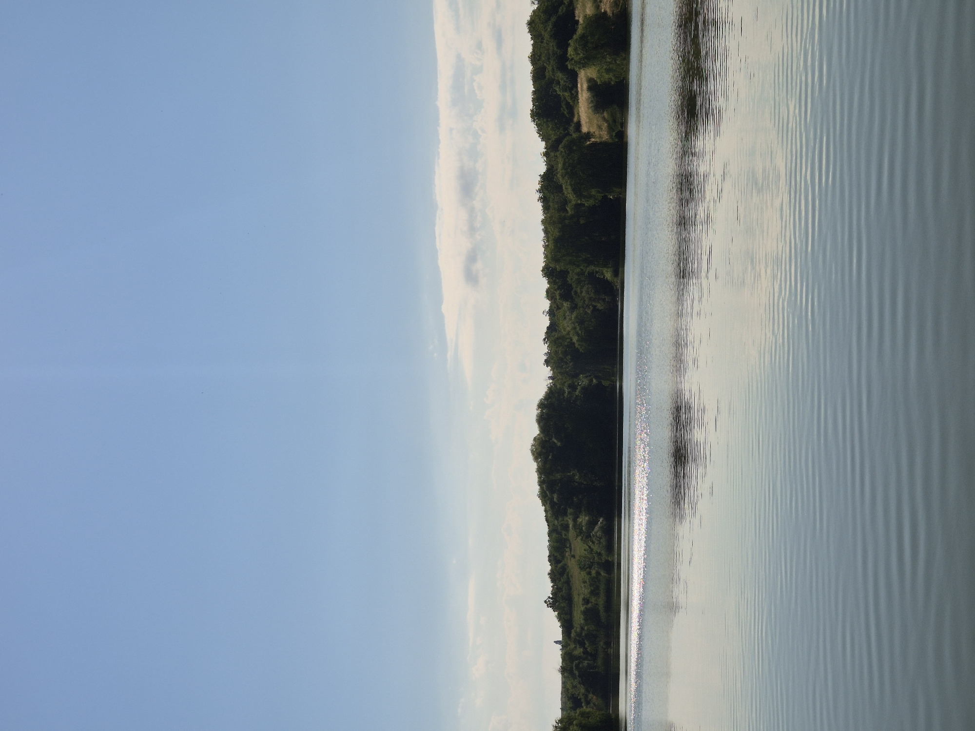 This is a wide shot of a tranquil lake on a clear day. The sky is a brilliant, cloudless blue, with a few faint wispy clouds near the horizon. The lake's surface is calm, with gentle ripples creating a textured pattern. Sunlight reflects off a section of the water, creating a shimmering effect. The far bank of the lake is lined with dense, green trees and shrubs, with a gently rolling, grassy hillside visible behind them. A small, solitary monument or structure can be seen on the crest of the hill. The overall impression is one of natural beauty and serenity. The location is the reservoir in the village of Sozonivka, Ukraine.