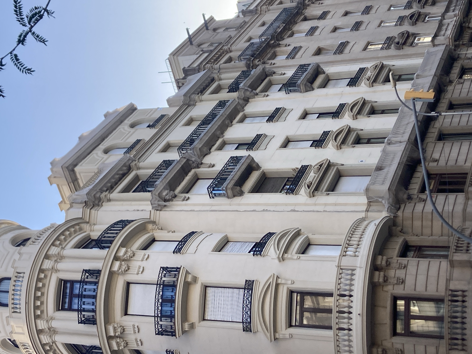 This media file captures a low-angle, upward view of an ornate, cream-colored building facade, likely in Barcelona, Spain, given the architectural style. The building features numerous windows, many with intricate black wrought-iron balconies or decorative window grilles. The architectural details are elaborate, with ornate moldings, sculpted elements above some windows, and rounded bay windows on the lower floors. A curved, balustrade-like structure is visible on the left, wrapping around a section of the building.
The scene appears to be set during the daytime, with a bright, clear sky visible at the top of the frame. A few dark branches from a tree are also visible against the sky in the upper left corner. A gray pole with a yellow rectangular object, possibly part of a street lamp or signage, extends vertically on the right side of the image. The building's facade is well-lit, suggesting sunny weather. No people are visible in the media. The overall impression is one of historical architecture bathed in daylight, highlighting the detailed craftsmanship of the building. The image does not contain any visible text.