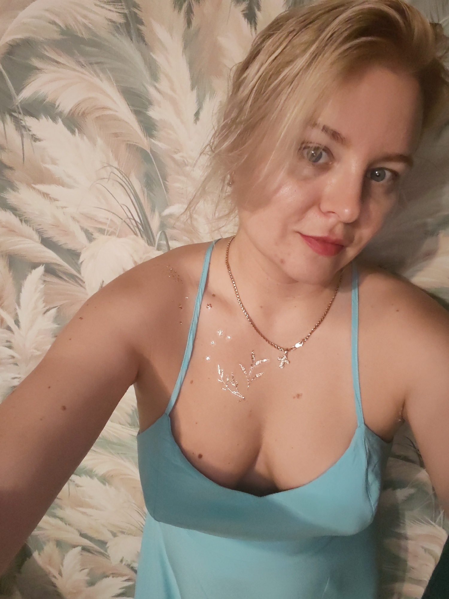 A young Caucasian woman with blonde hair and blue eyes is pictured lying down against a wall with a light-colored, fluffy pattern. She is wearing a light blue dress with thin straps and a necklace with a small pendant. Her left shoulder and upper chest are adorned with temporary silver glitter tattoos in the shape of stars and a sprig. The image appears to have been taken indoors, possibly in a bedroom in Izhevsk, Russia. The lighting is soft, creating a slightly diffused effect.