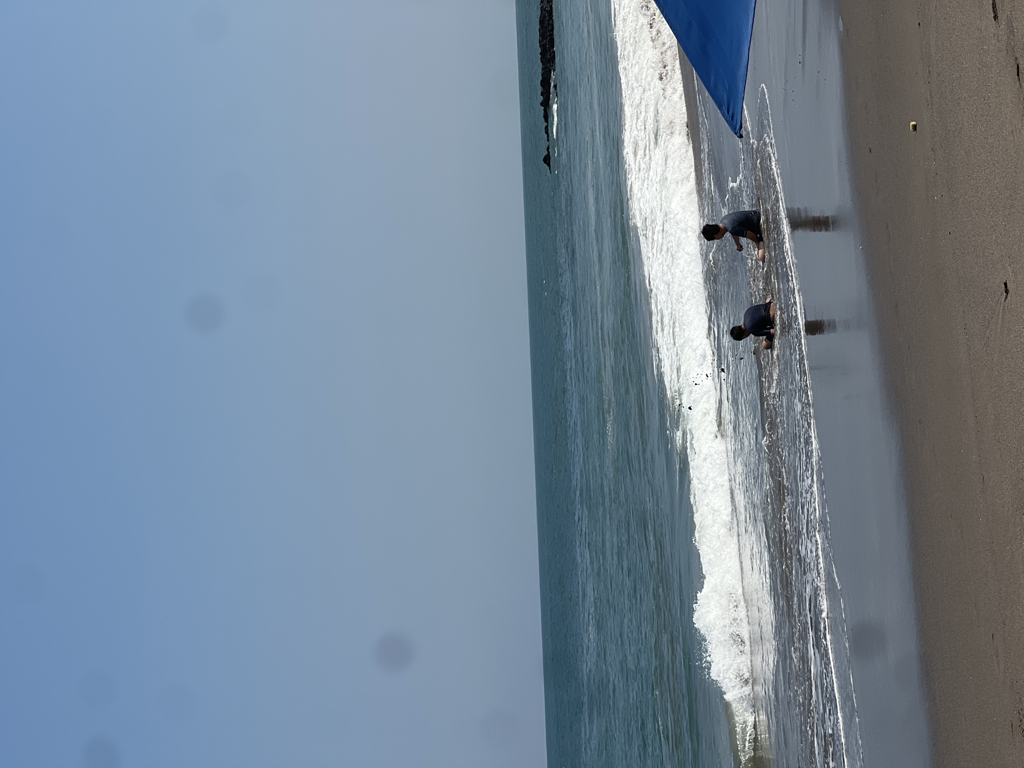 Two young boys are sitting in the shallow water at the edge of a dark sand beach. The boy on the left is closer to the water's edge and appears to be playing. The boy on the right is slightly further back and is also sitting in the water. A blue umbrella is partially visible on the right side of the frame, providing shade. The ocean is calm with gentle waves breaking on the shore. In the background, a rocky outcropping juts out into the water. The sky is clear and blue, with no clouds visible. The scene appears to be set in a tropical location, likely on the coast of North Kuta, Indonesia, given the dark sand beach and clear blue sky.