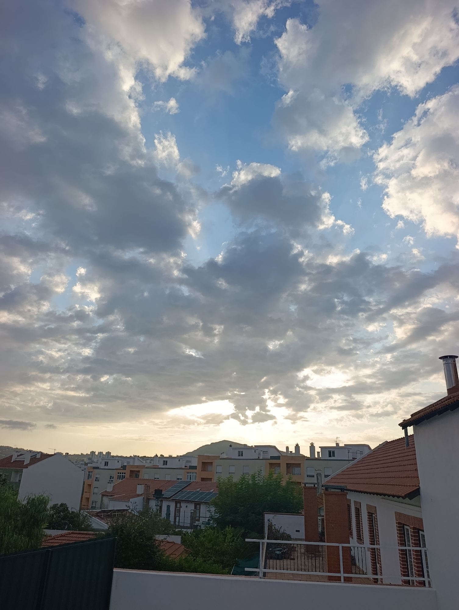 The image captures a view of the cityscape of Setúbal, Portugal, under a dramatic sky filled with clouds. In the foreground, the roof of a white building with a tiled roof and a chimney is visible on the right. The middle ground features a dense collection of urban architecture, including buildings with terracotta tiled roofs, white facades, and some with solar panels. Green trees and foliage are interspersed among the buildings. The background shows a line of residential buildings stretching across the horizon, with a distant hill or mountain range under a hazy, overcast sky. The sky itself is a mix of light blue patches and heavy, gray clouds, with the sun partially obscured, creating a soft, diffused light. The overall impression is of an urban environment at dusk or during a cloudy day.
