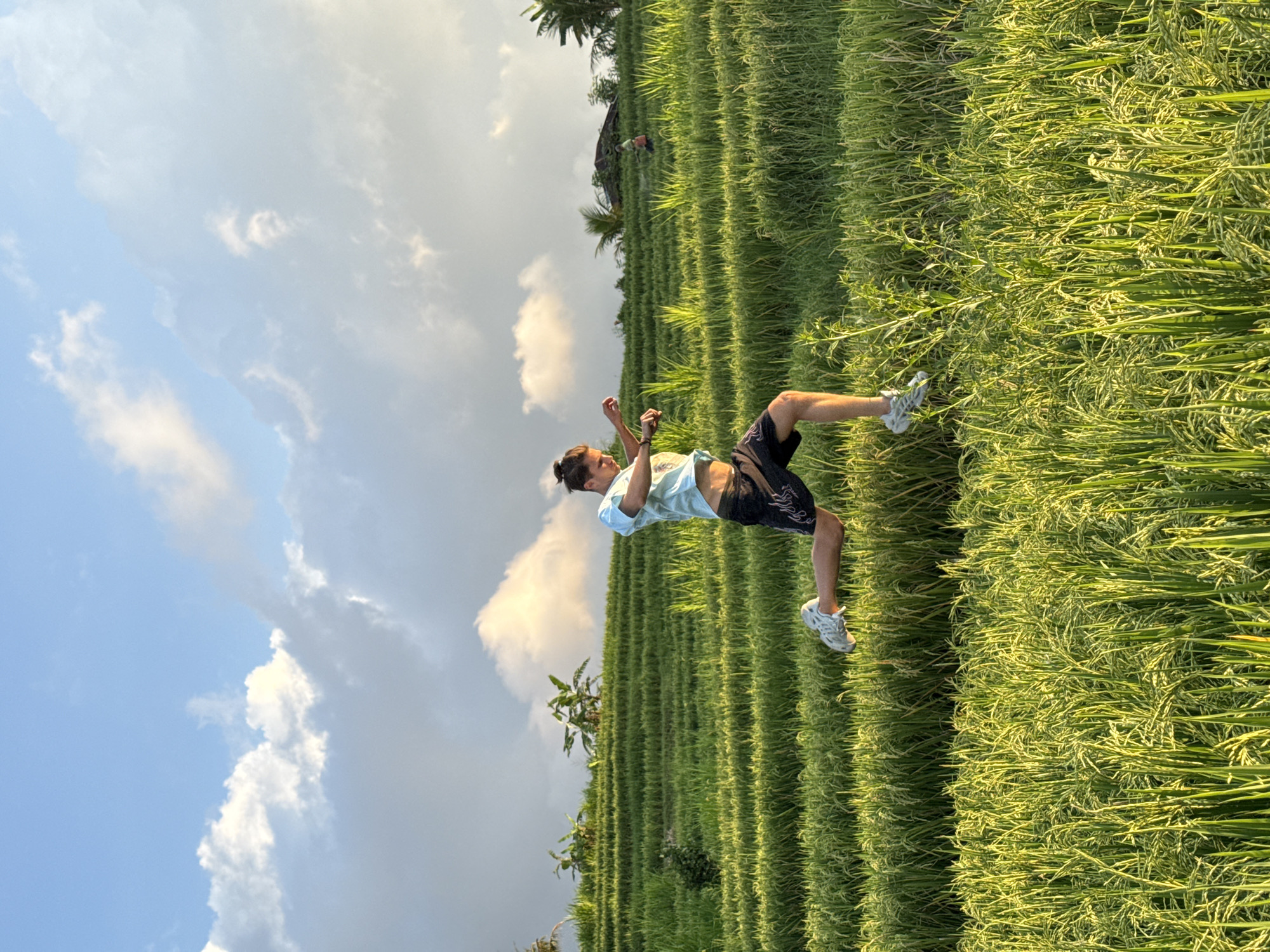 A man is captured mid-air, positioned at an approximate 45-degree angle against a backdrop of a rice field. The location is Padangbulia, Indonesia. The man wears light-colored t-shirt, dark shorts, and light-colored sneakers. He is in the middle of a backflip. The rice field features rows of green stalks. The sky behind the rice field is filled with white clouds and a gradient of light blue to a near-white sky.