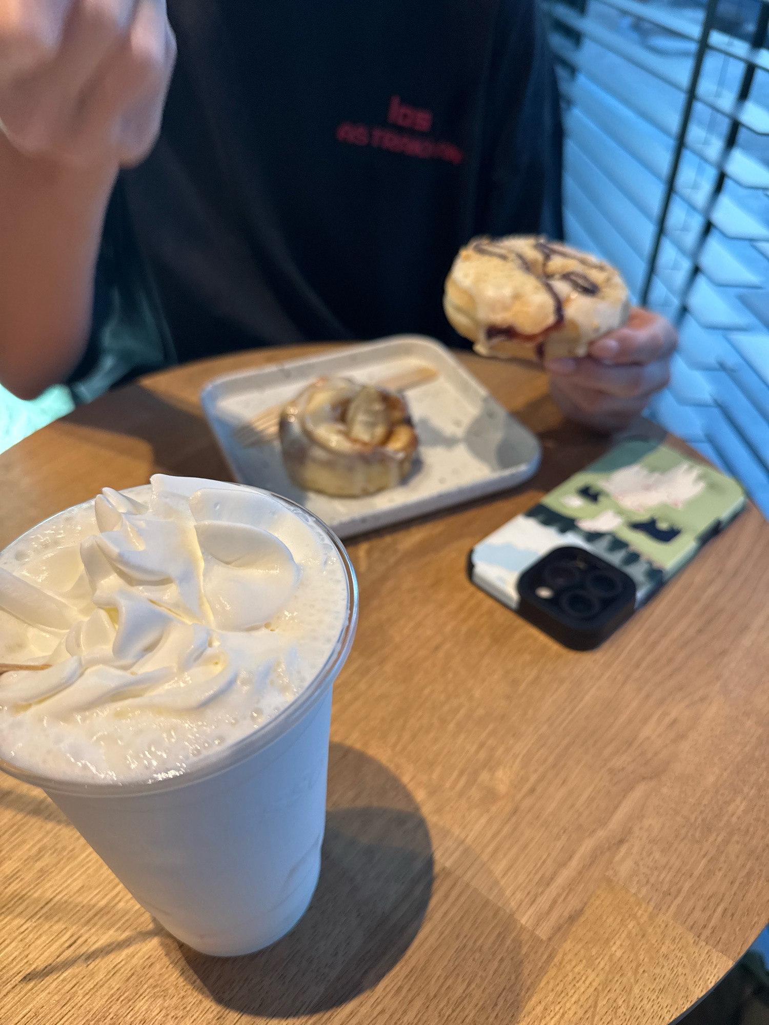A person's hands are holding a donut with chocolate drizzle and a bite taken out of it, above a table. To the left, a white plastic cup filled with a white, frothy drink topped with whipped cream is on the table. To the right, a smartphone with a green and white camouflage case, showing a camera lens assembly, rests on the table. In the background, a white plate with a cinnamon roll on it is visible, along with part of a person wearing a black t-shirt with red lettering. The scene appears to be set in an indoor cafe with window blinds in the background. The image does not provide any specific geographical markers to indicate the city of Antalya, Turkey.