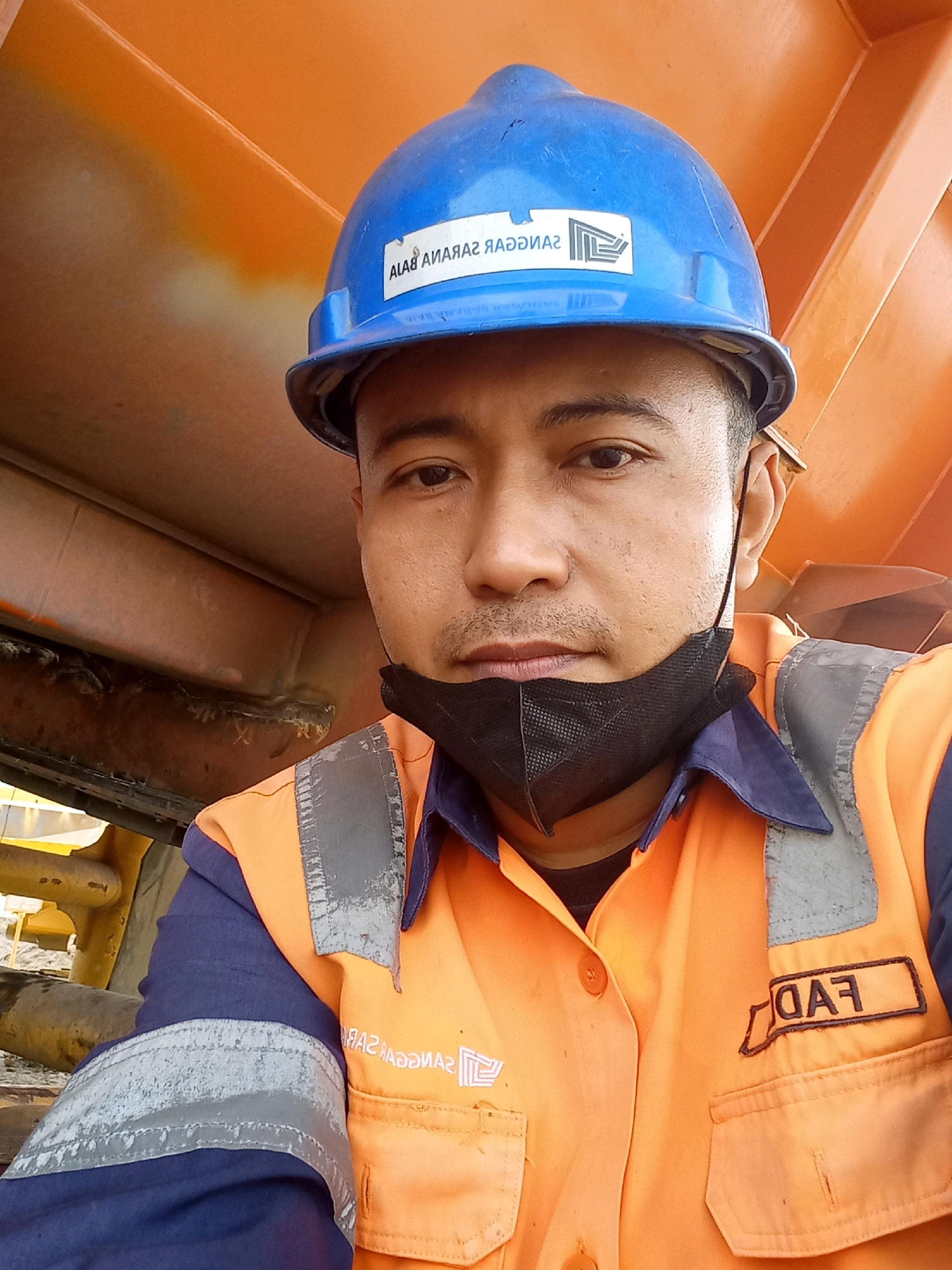 A man wearing a blue hard hat and an orange high-visibility vest takes a selfie outdoors. The vest is over a dark blue collared shirt. He has a black face mask on, pulled down below his nose. The background is predominantly an orange structure, possibly a vehicle or building. Text and a logo are visible on the hard hat and the vest. The logo on the hard hat appears to be a stylized building or structure. The text on the vest reads "FAD". The man has short dark hair and is looking directly at the camera. The overall scene suggests an industrial or construction environment.