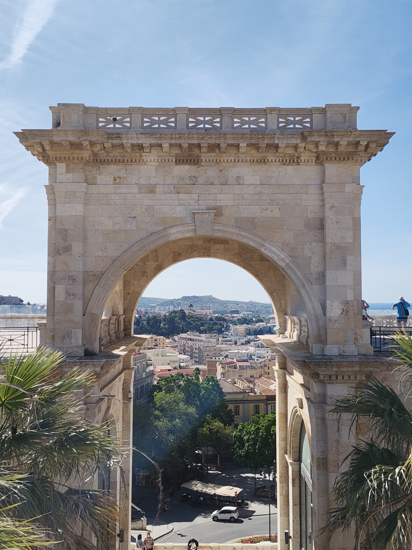 The image depicts the Bastione di Saint Remy in Cagliari, Italy, framed from a slightly low angle through its arched opening. Constructed from light-colored stone, the Bastione features a decorative parapet at the top. The archway provides a view of the city below, with buildings visible in the distance, reaching up to a hilly horizon. Trees partially obscure the view of a street just beneath the Bastione, where pedestrians walk and a white car drives. Two people stand on the right side of the Bastione, overlooking the city. Palm trees frame the left and right sides of the view. The sky is blue with scattered clouds.